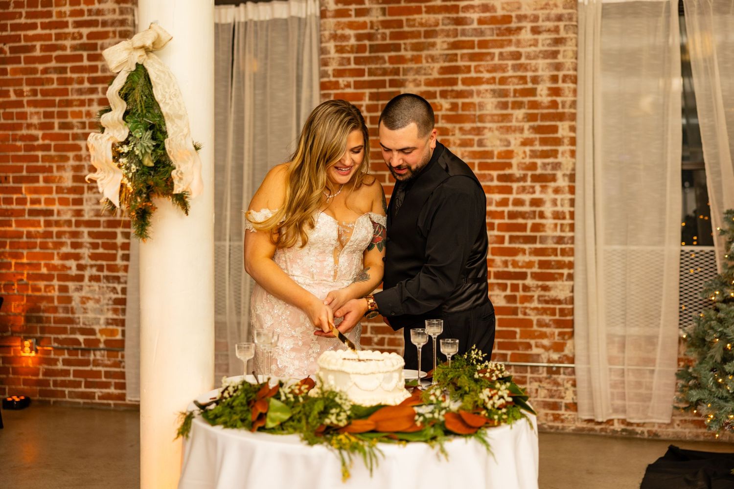 Couple cutting their wedding cake together beside a decorated brick wall.