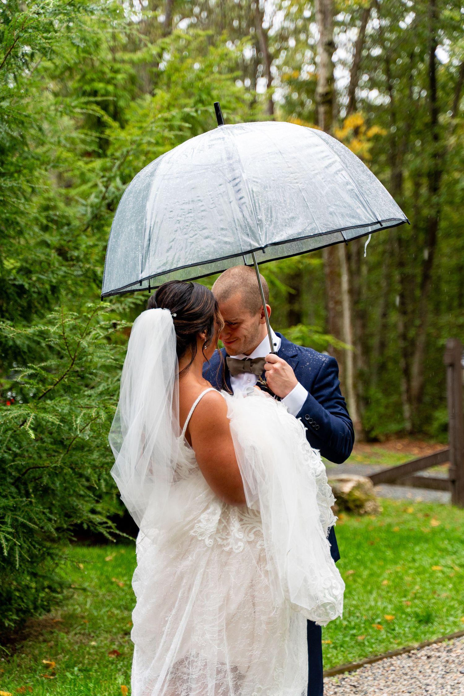 Romantic moment between newlyweds under clear umbrella in lush garden setting.