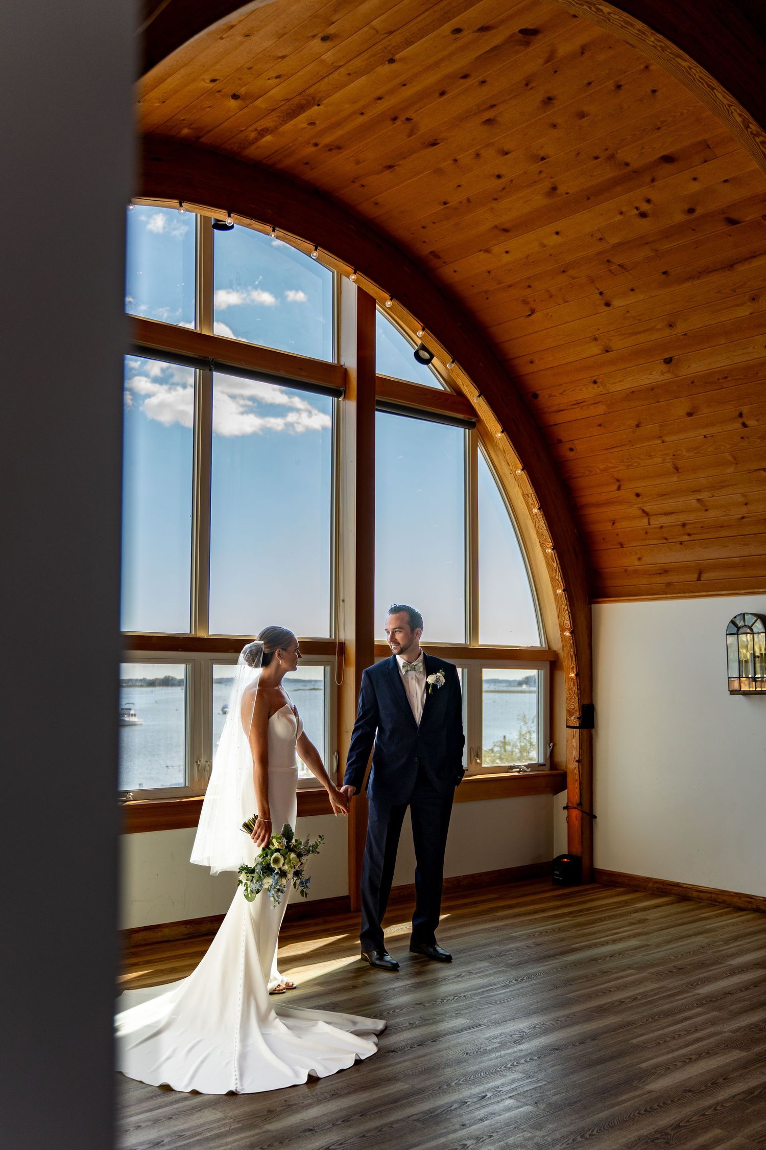 Wedding couple stands in sunlit room with arched wooden ceiling and large windows.