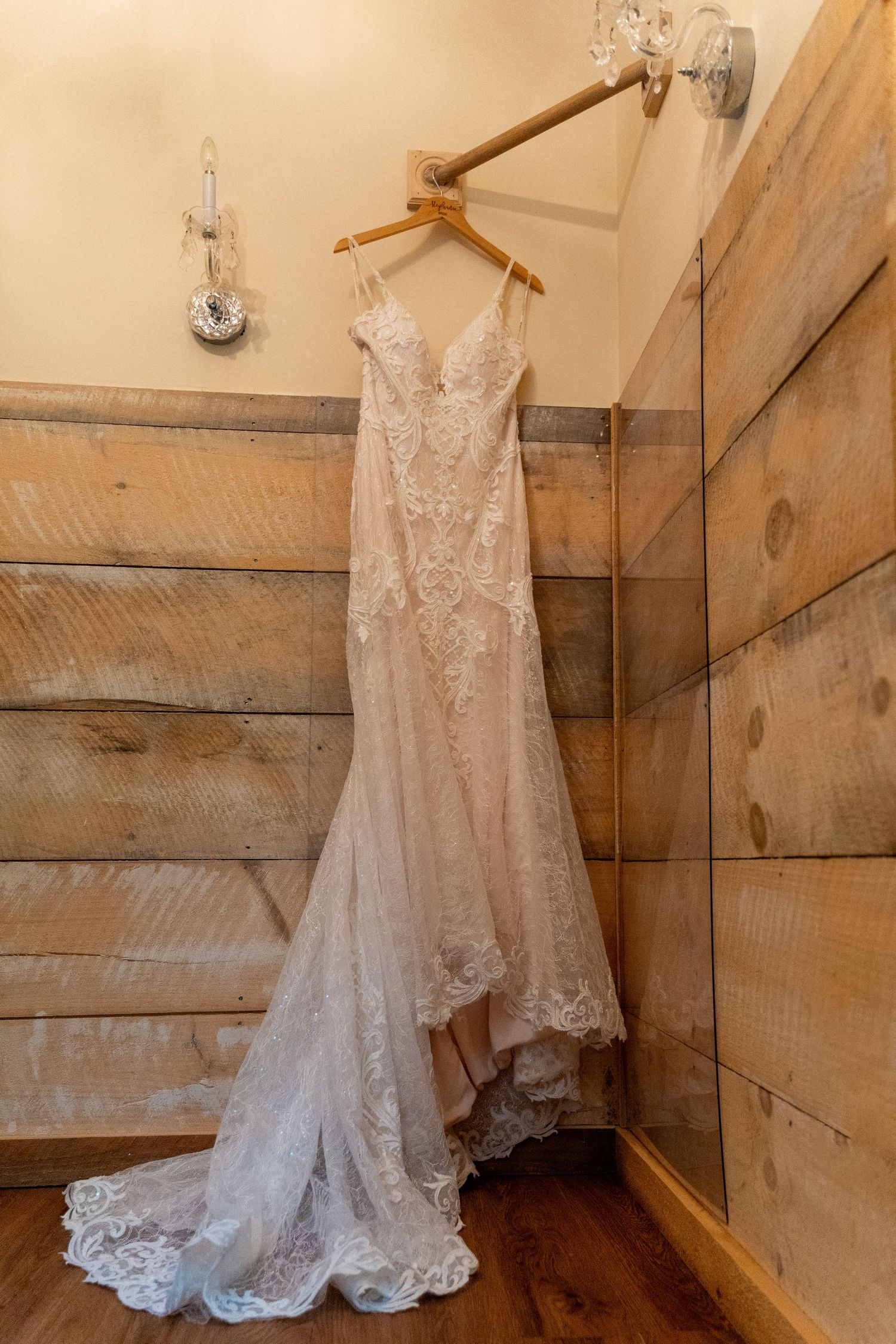 Elegant lace wedding dress hangs on wooden wall with delicate train flowing down.