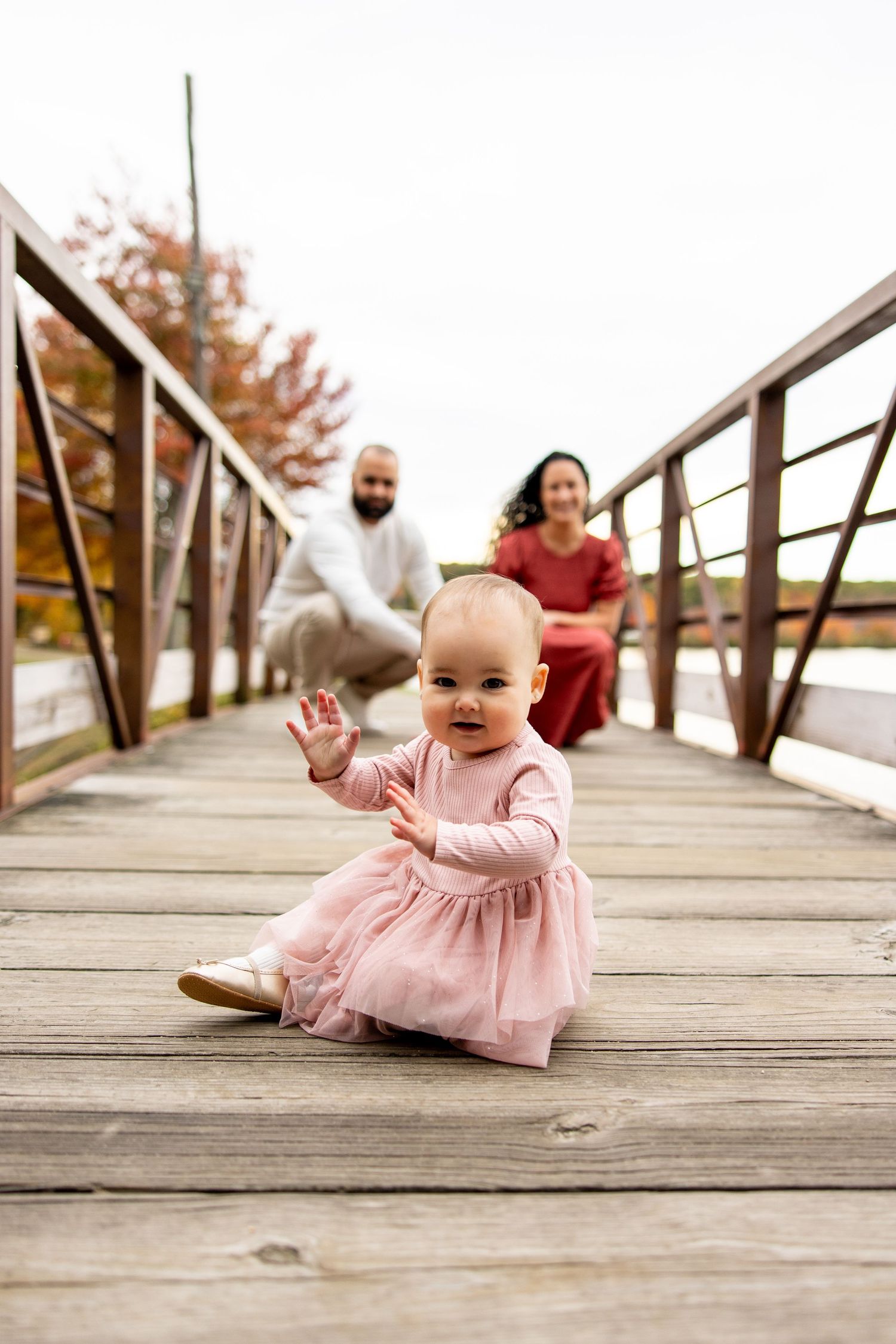 Baby in pink dress sits on wooden bridge while parents watch from behind.