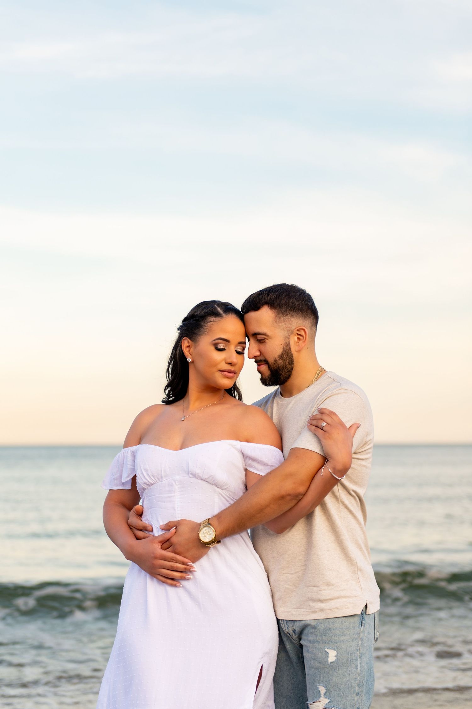 A romantic couple embraces on the beach during golden hour with ocean waves behind them.