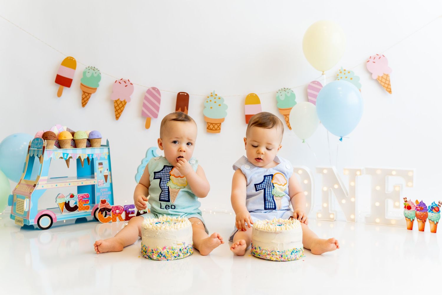 Twin babies in matching outfits enjoying their first birthday cakes with ice cream party decorations.