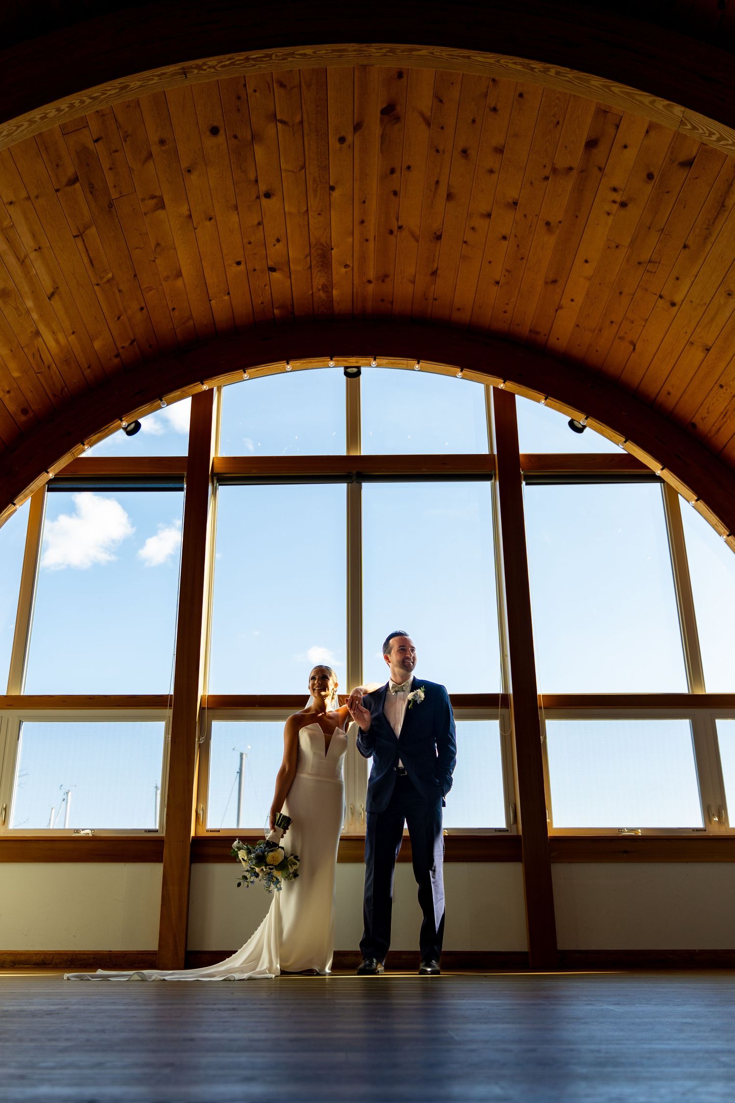 A couple stands silhouetted against large arched windows with wooden beams in an elegant venue.