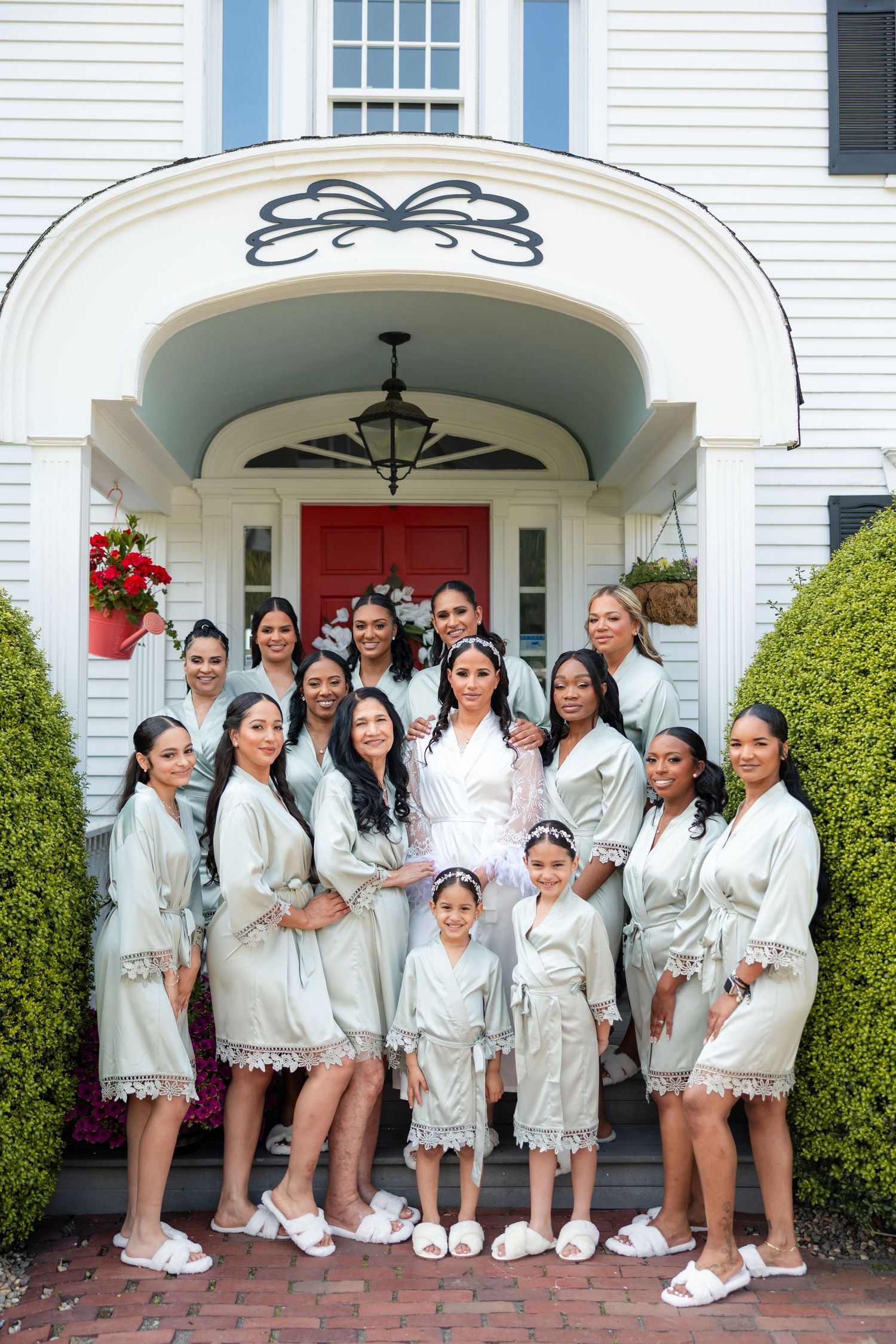 A group in matching white satin robes poses on the steps of a white house with a bright red door and arched entryway.