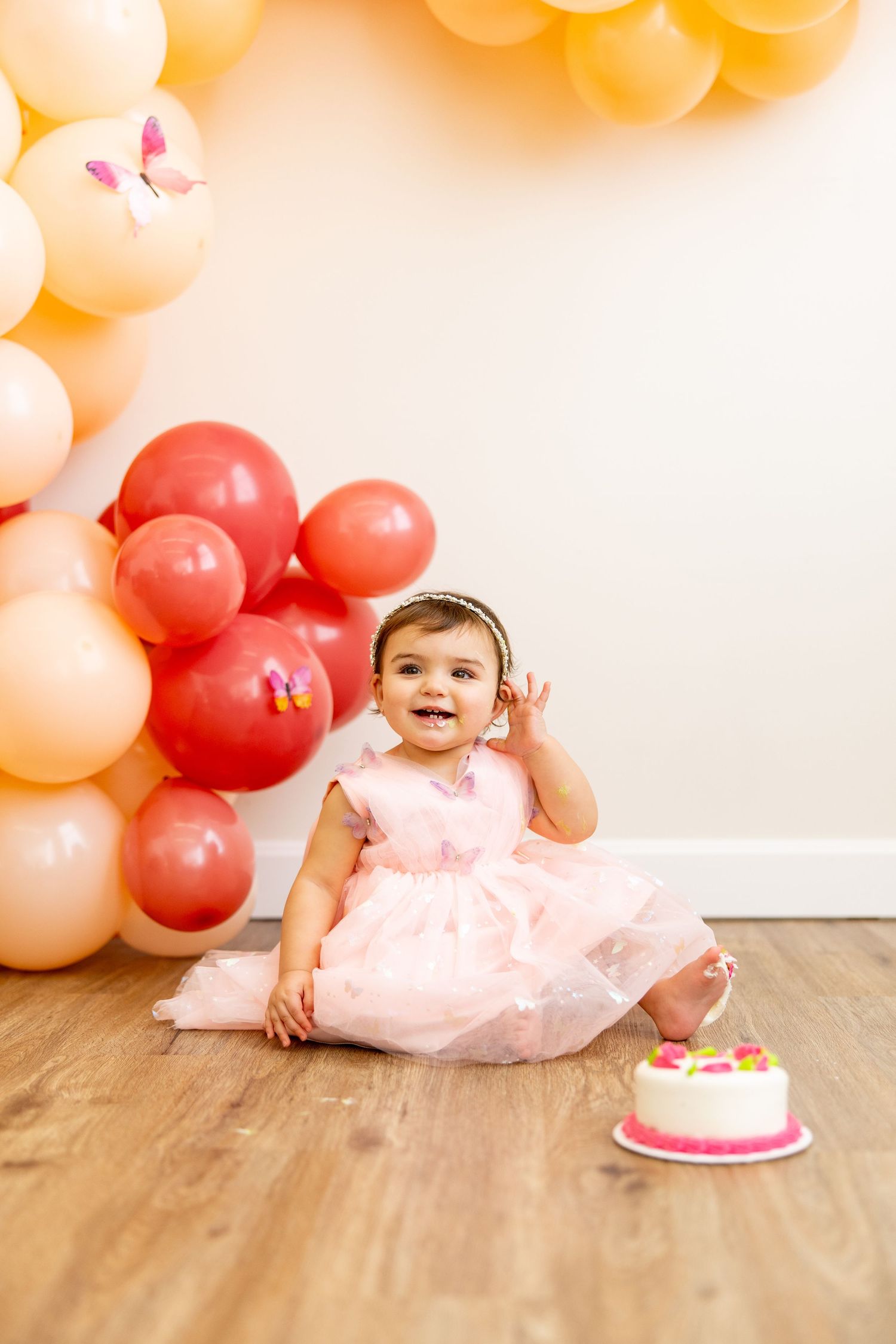 Baby in pink dress sitting by small cake with balloon arch backdrop.