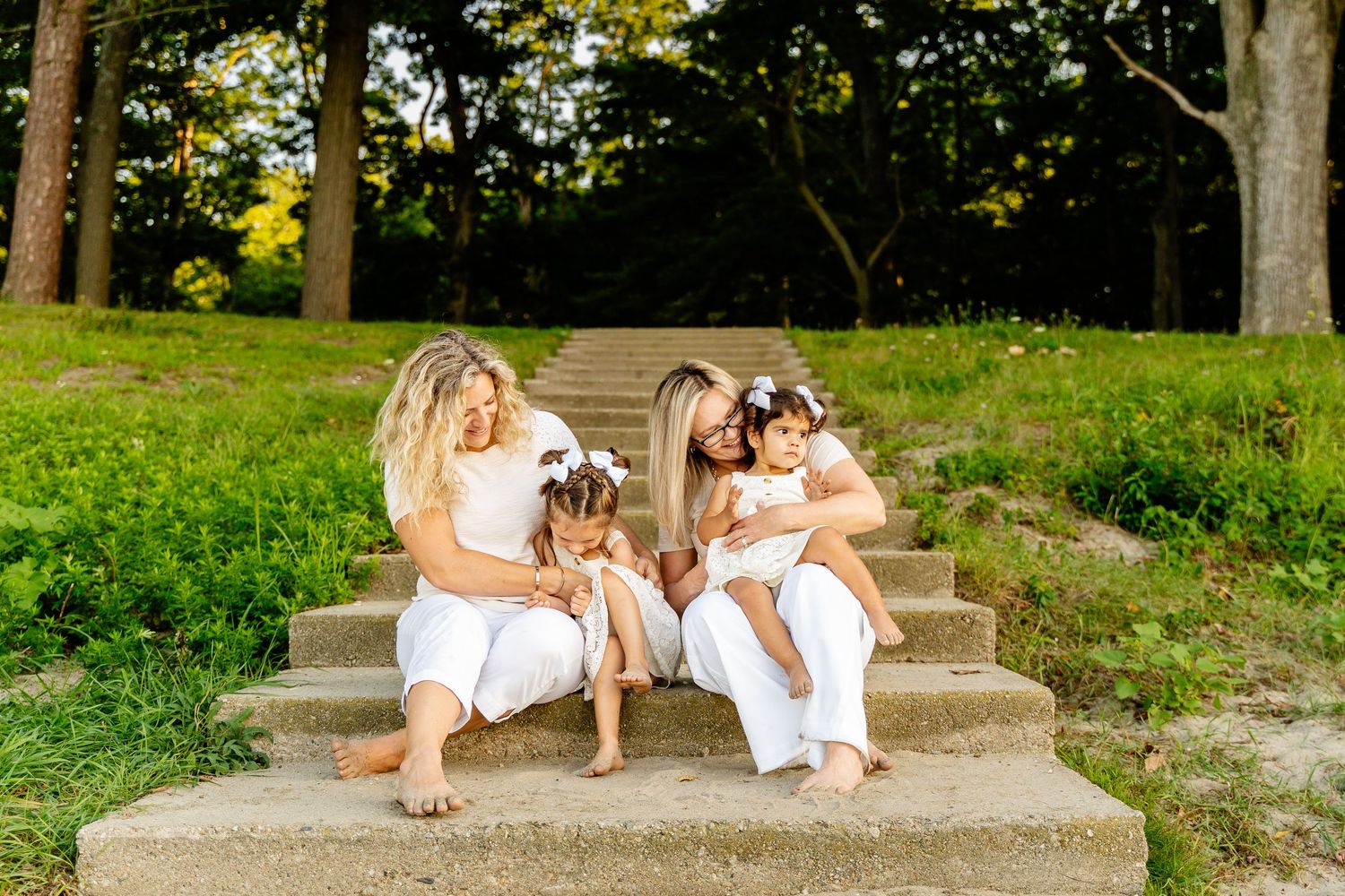 Family group sitting on concrete steps surrounded by lush green trees at sunset.