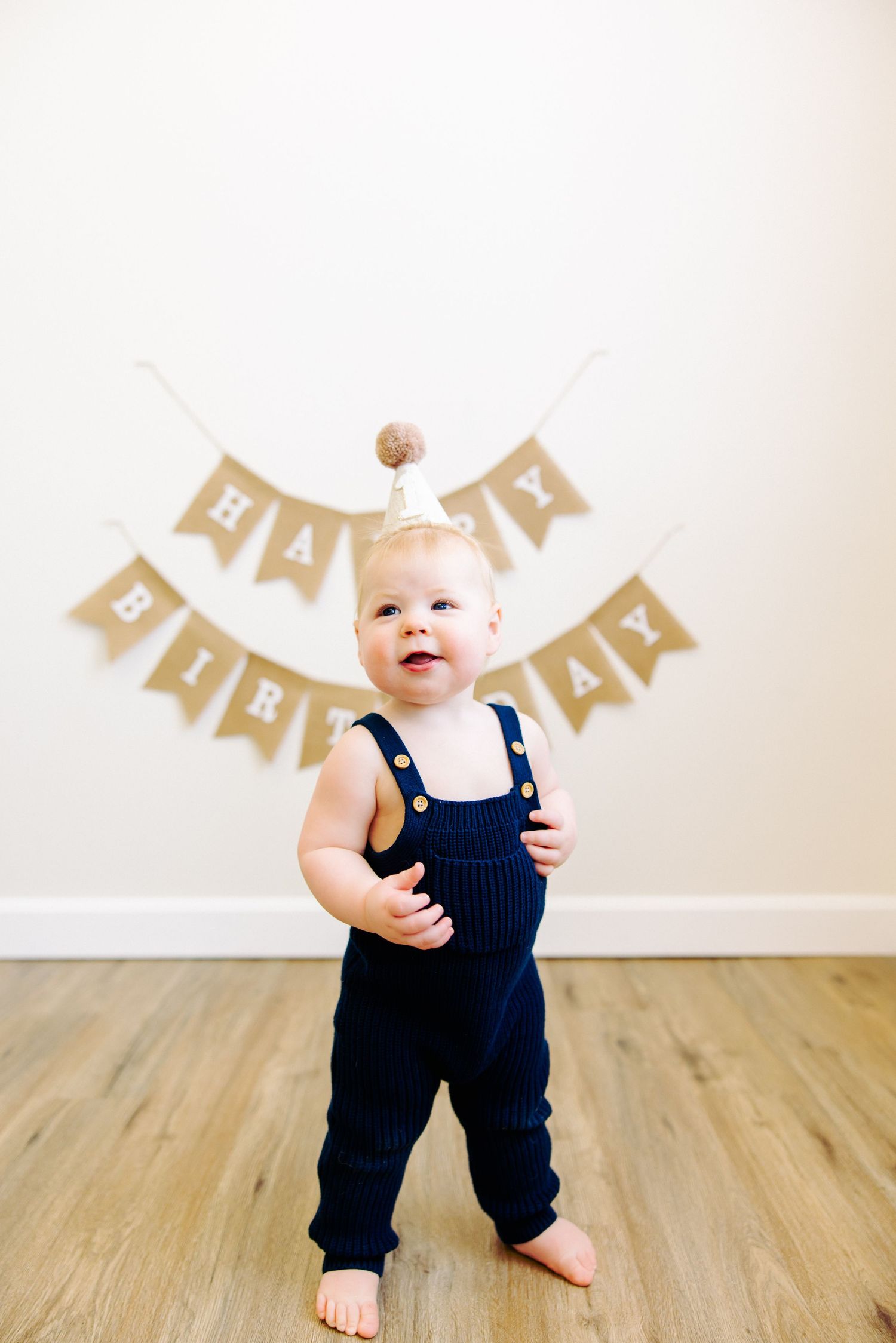 Smiling baby in navy overalls standing beneath burlap birthday banner.