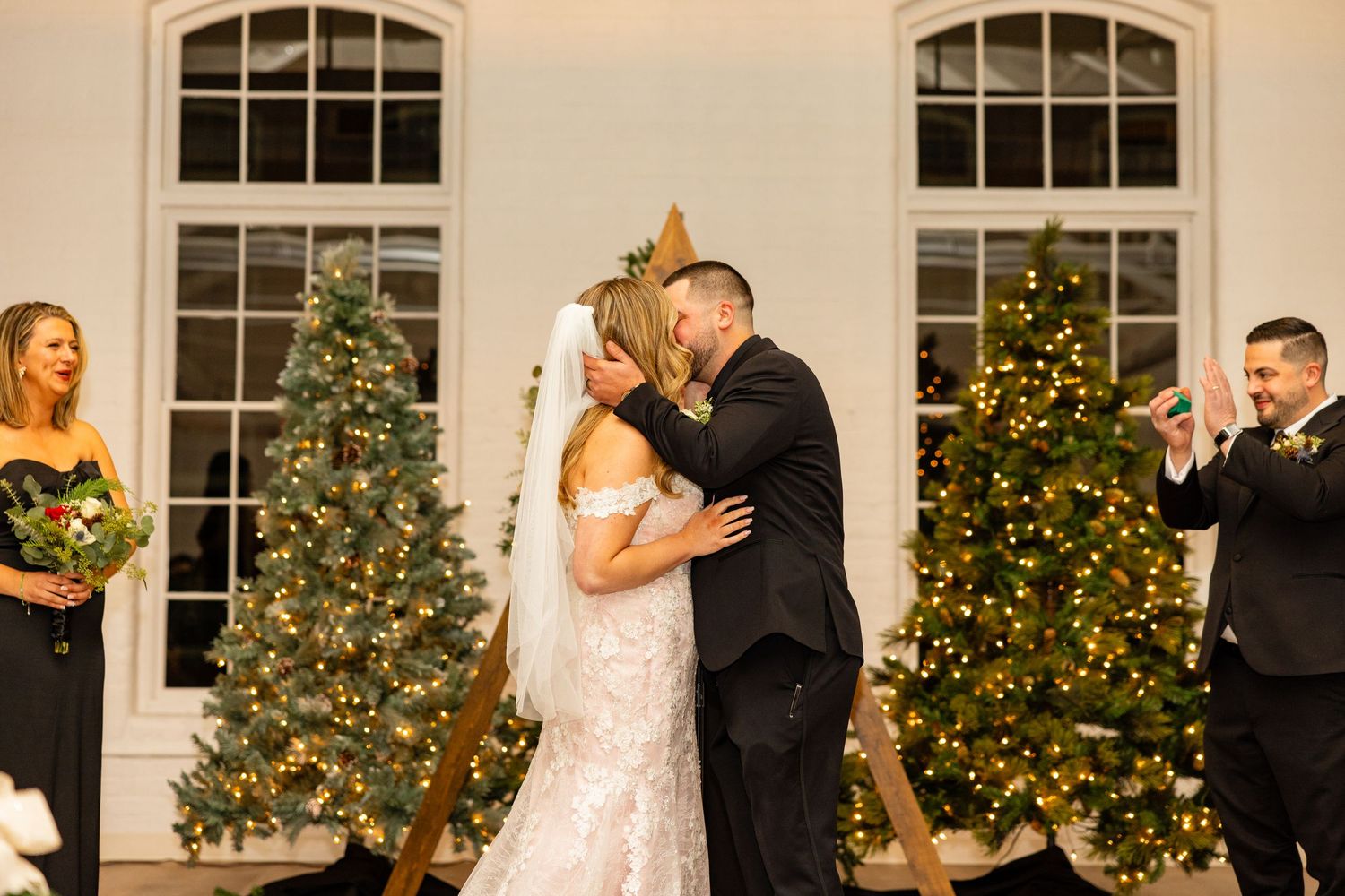 A couple shares their first kiss as newlyweds with decorated Christmas trees in the background.