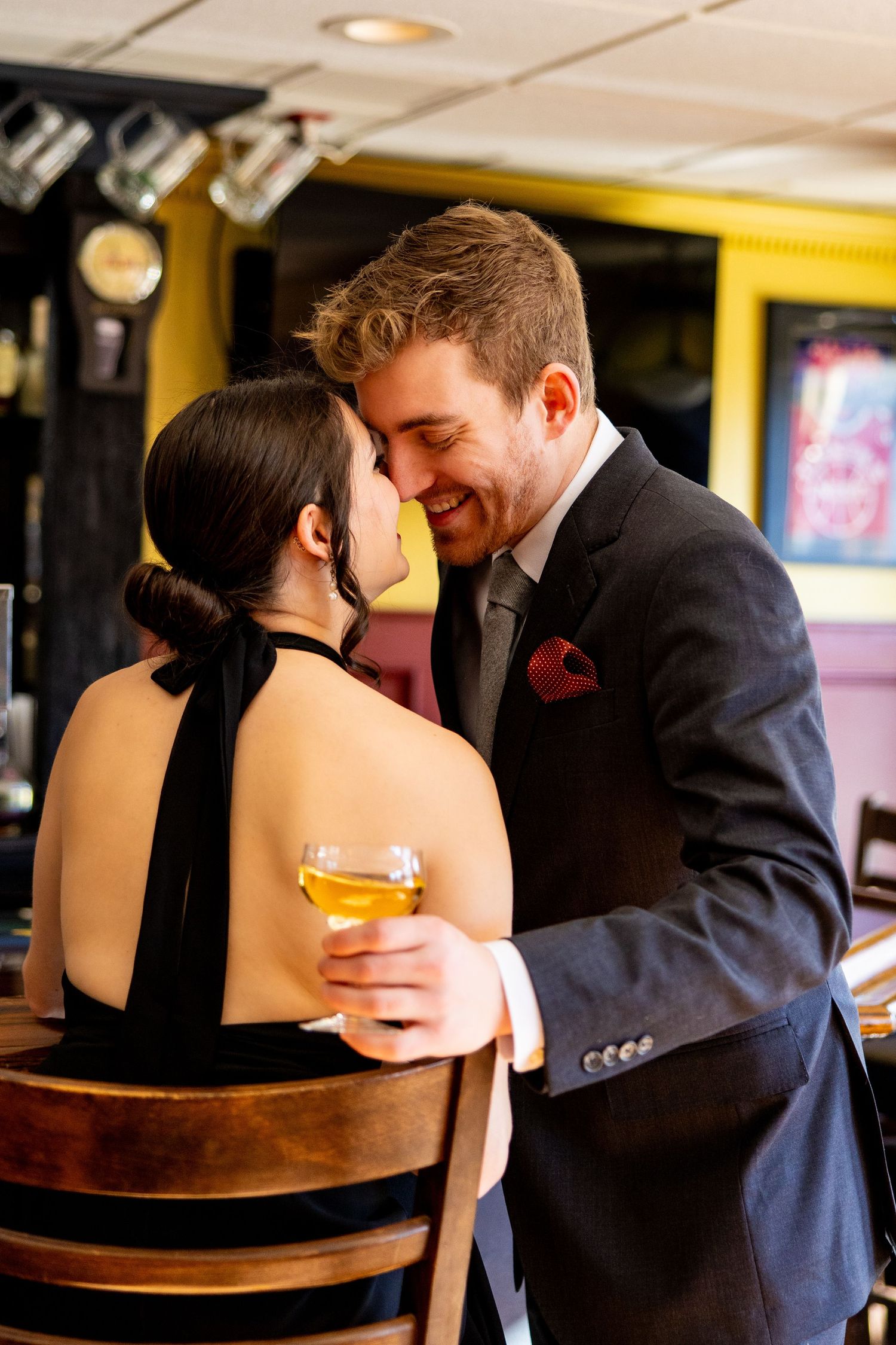A romantic couple shares an intimate moment at a bar while holding a cocktail glass.