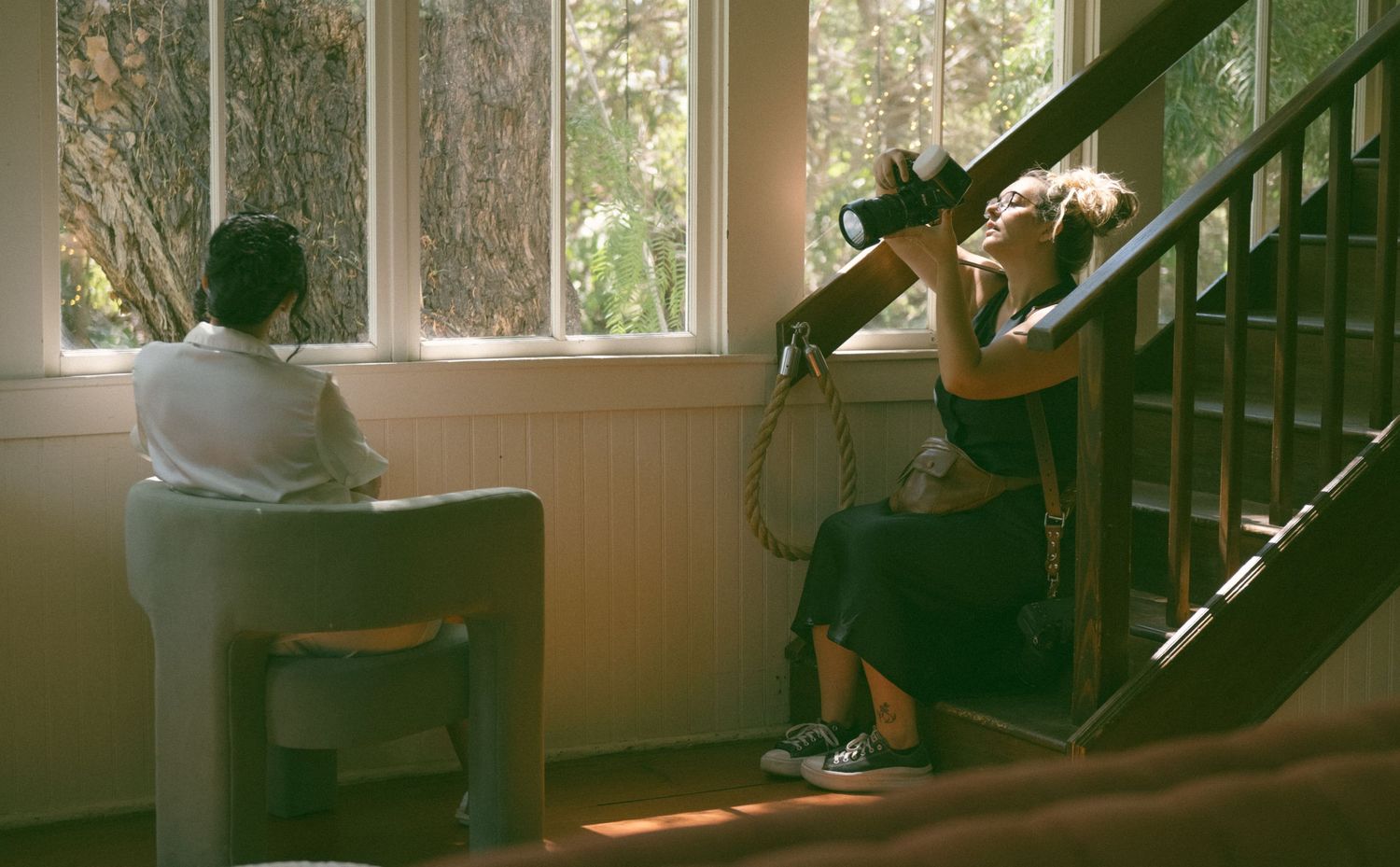 Two people sitting separately in a sunlit room with stairs and windows.