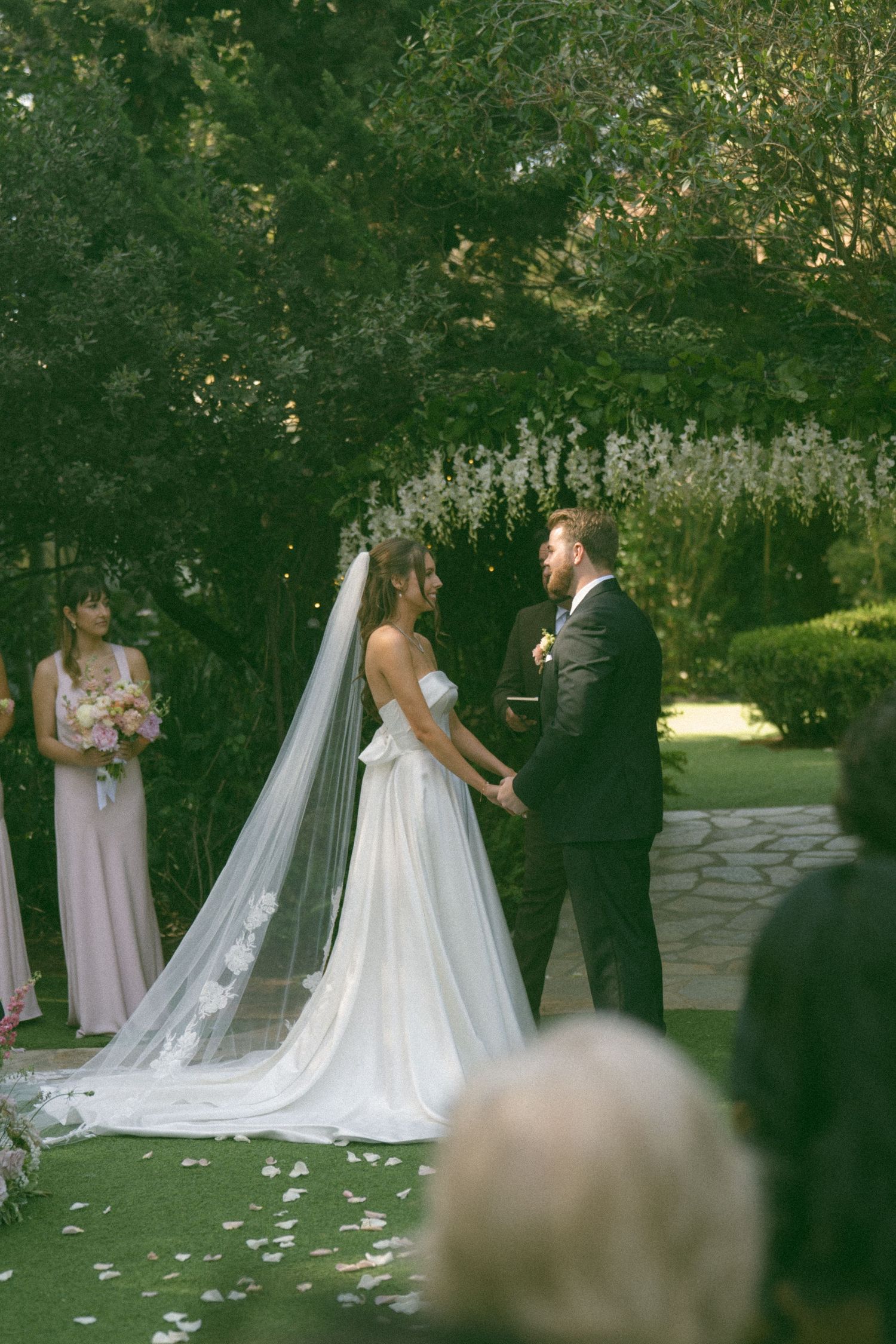 A wedding ceremony takes place in a garden setting with a long flowing veil and floral backdrop.