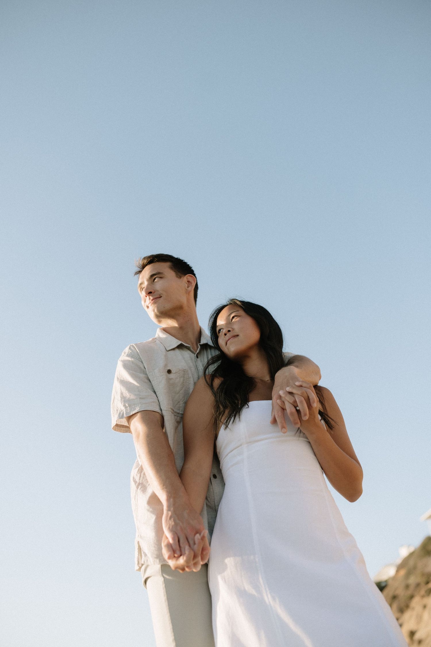 A romantic couple in white outfits poses against a clear blue sky near coastal cliffs.