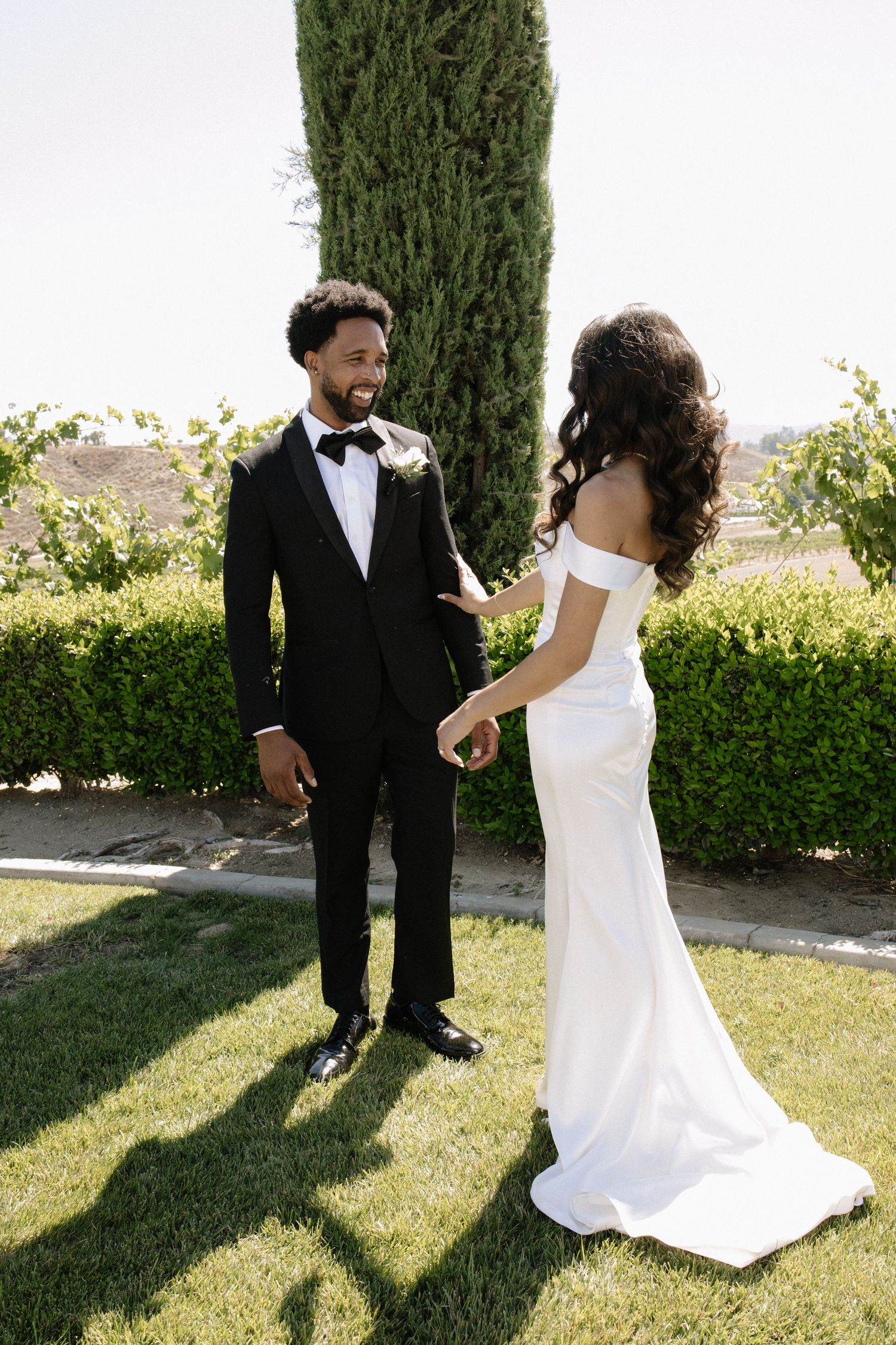 A couple in formal wedding attire poses playfully on a sunny lawn with cypress trees in the background.