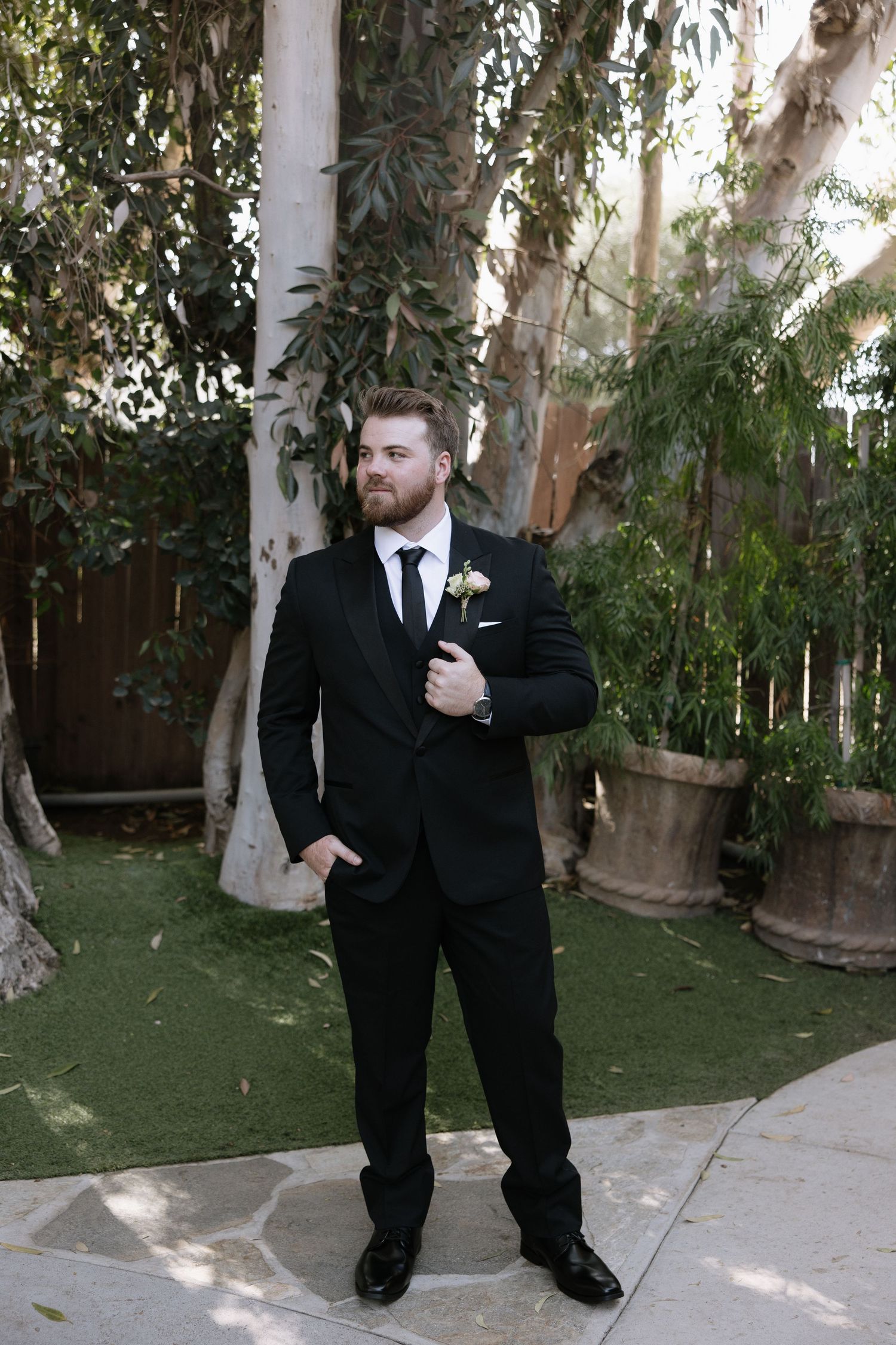 A formal portrait of a groom in a black suit standing against a garden backdrop.
