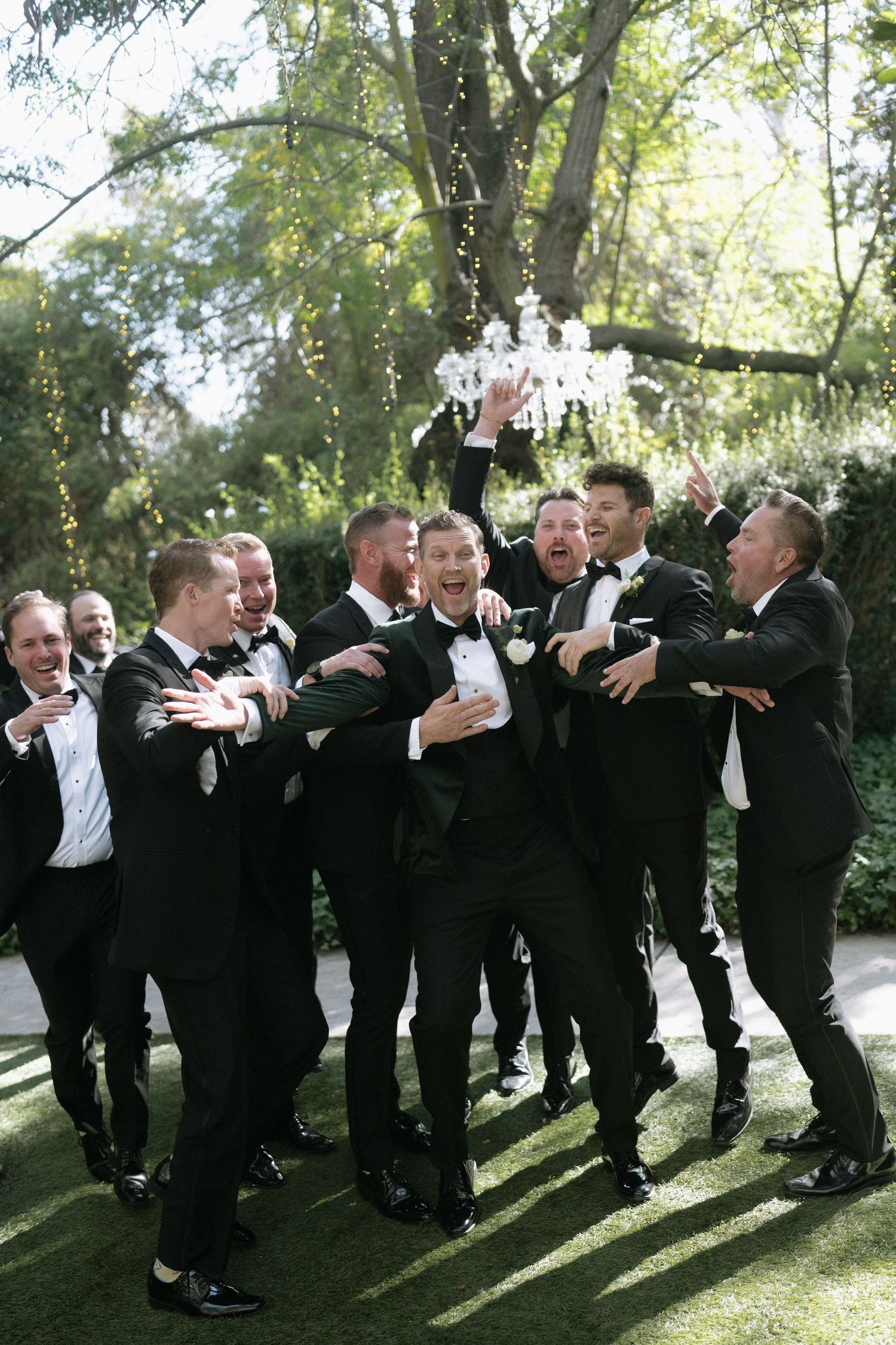 Groomsmen in black tuxedos pose playfully outdoors.
