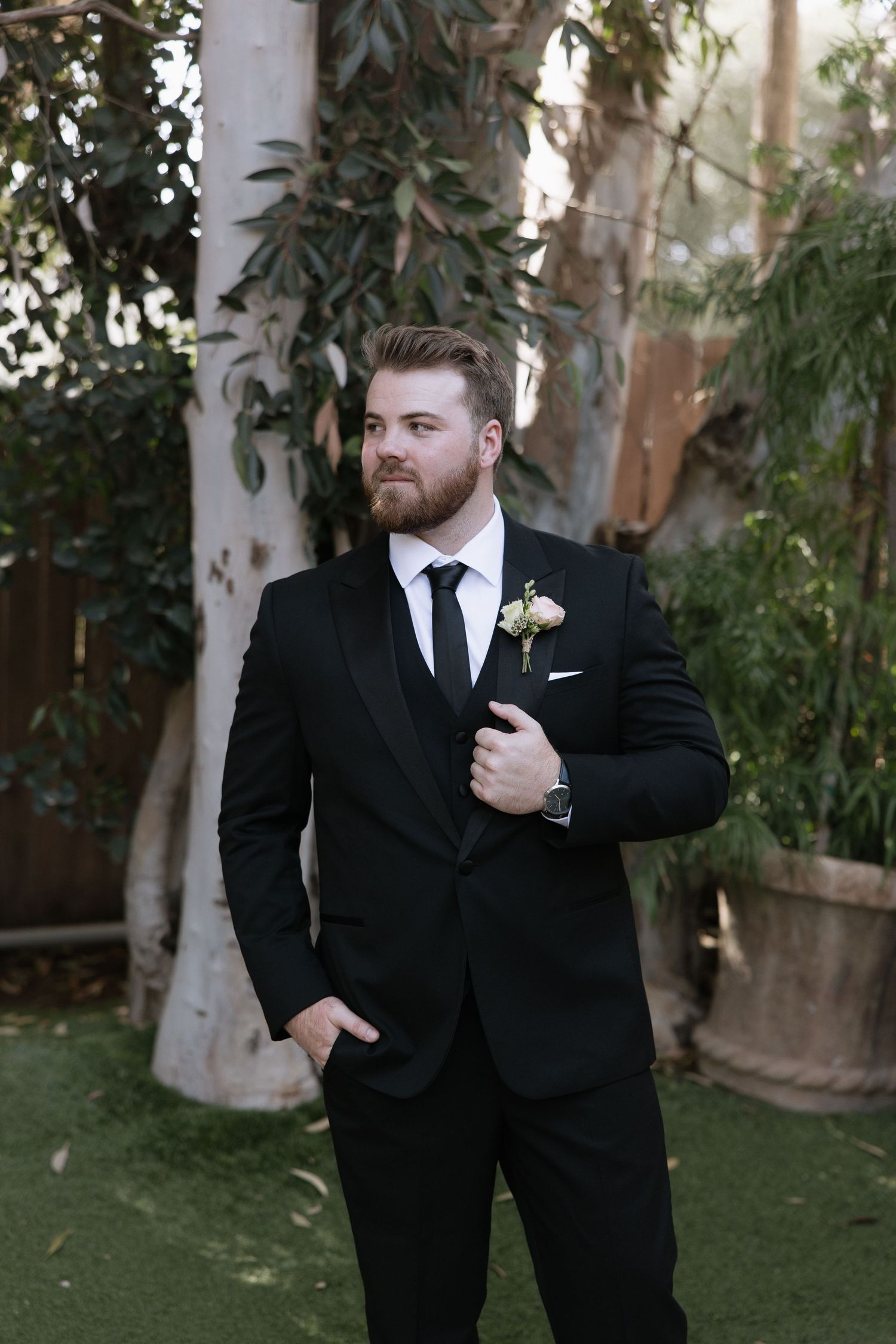 A groom in a classic black suit poses in front of eucalyptus trees.