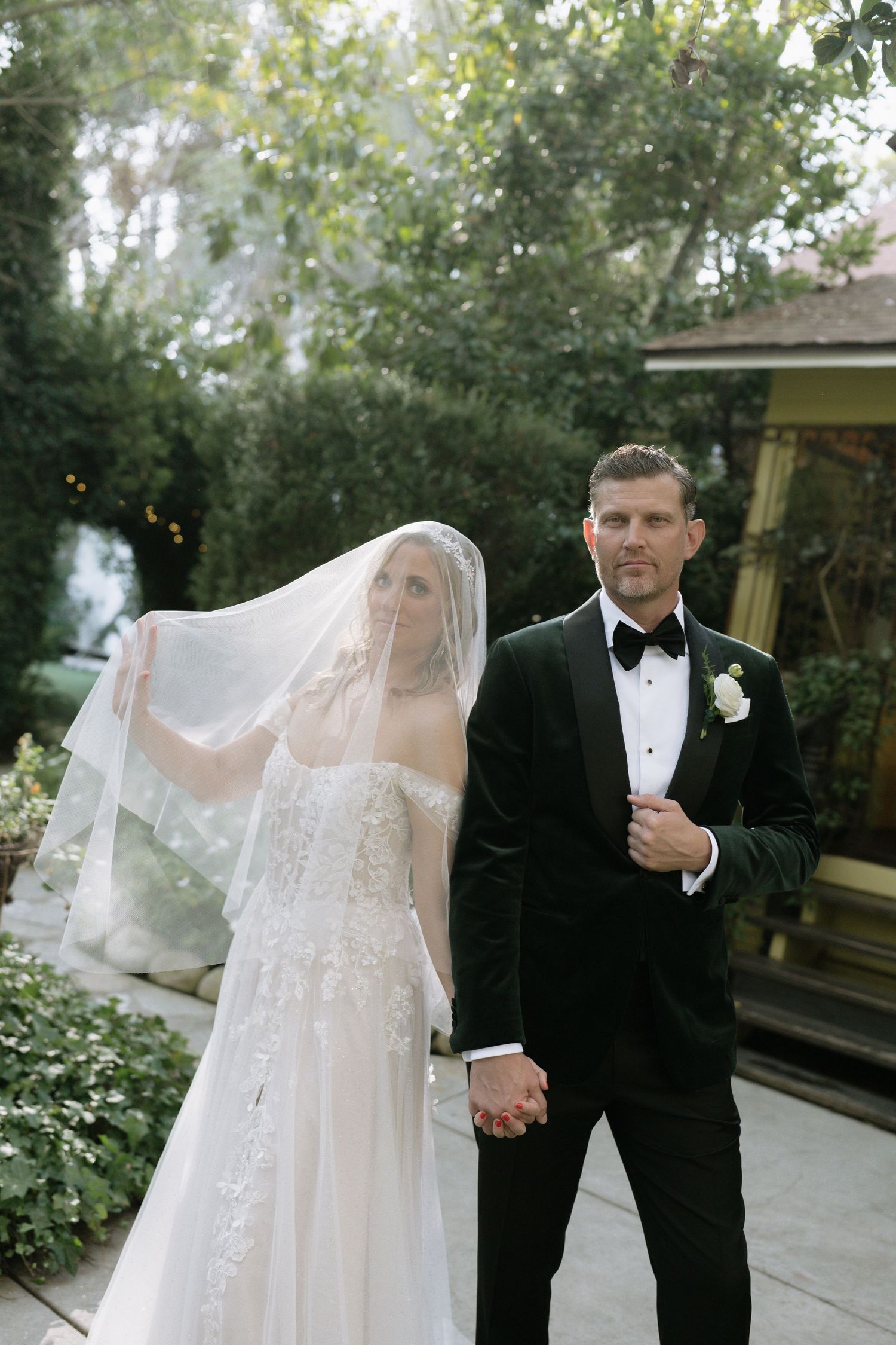 A romantic wedding couple shares intimate moments in a garden setting with a flowing veil and classic black tuxedo.