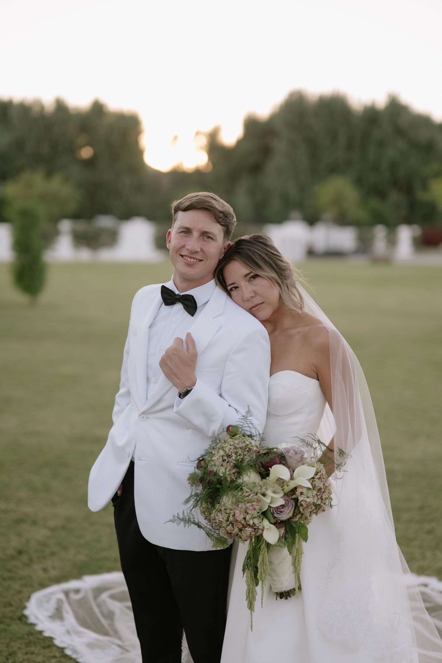 Newlywed couple in white formal attire posing in garden at sunset.