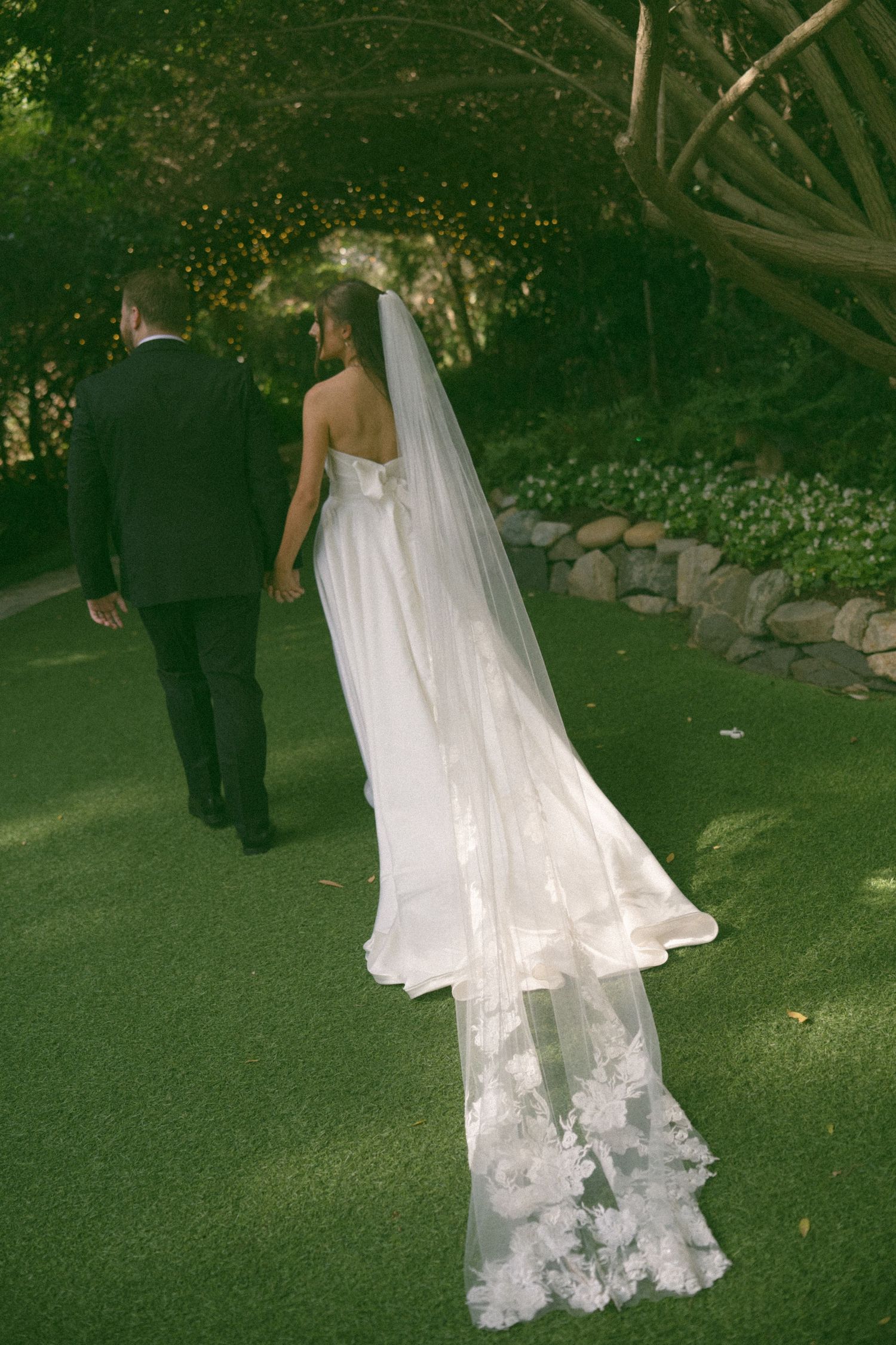 A couple walks along a garden path with a long flowing lace veil trailing behind.