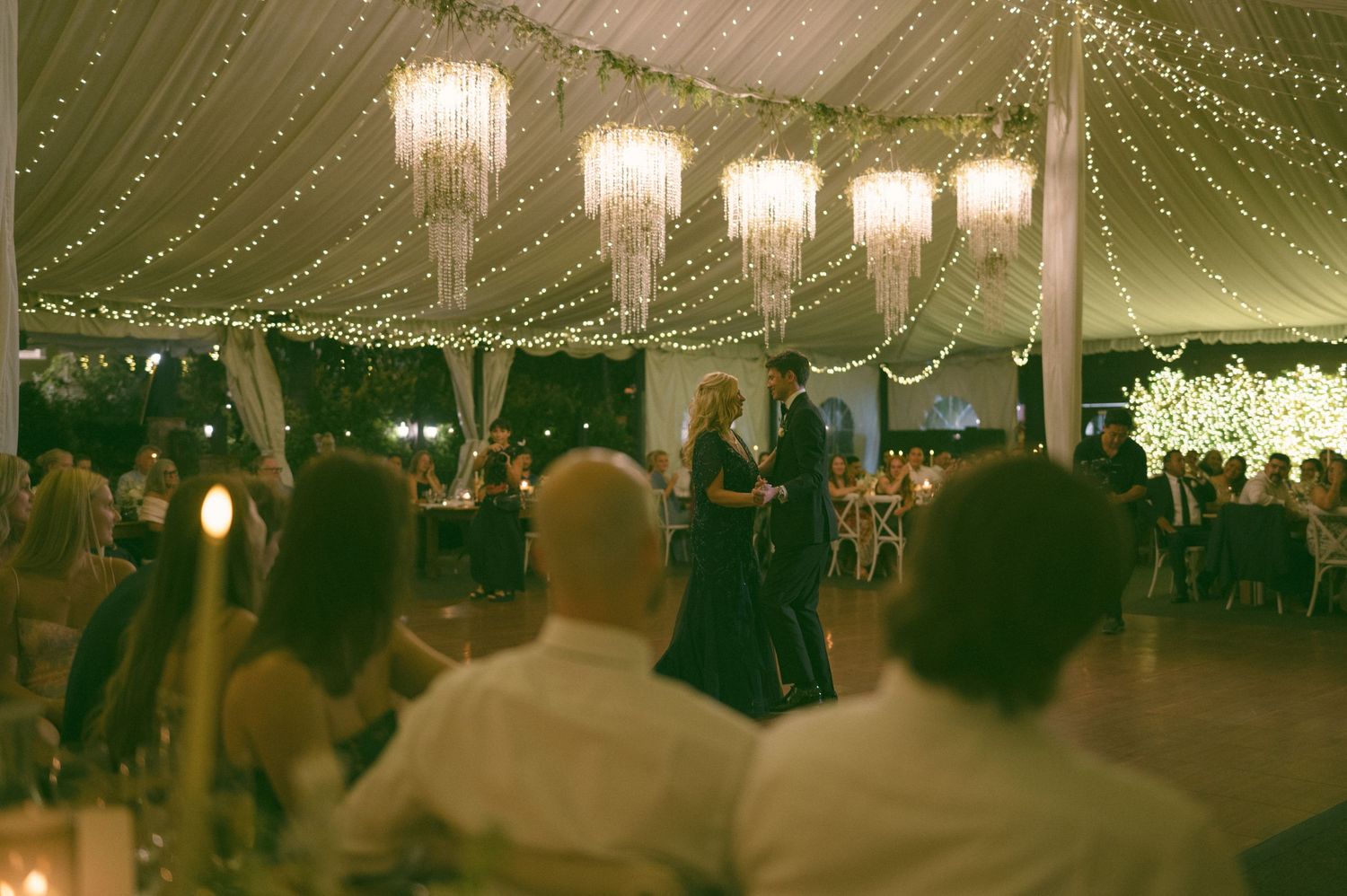 Wedding reception in an elegant tent with crystal chandeliers and string lights.