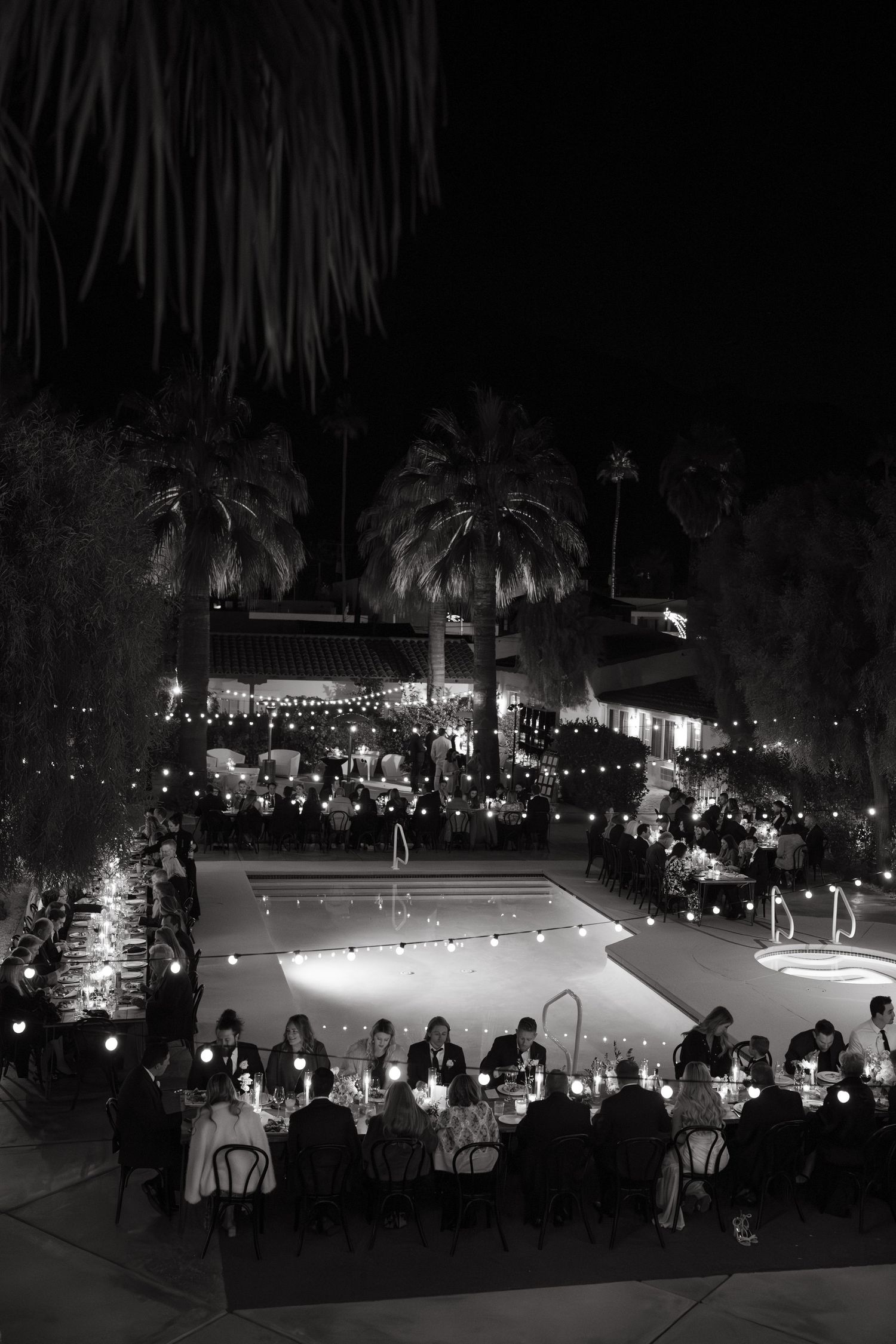 An elegant outdoor evening reception with string lights illuminating tables around a swimming pool.