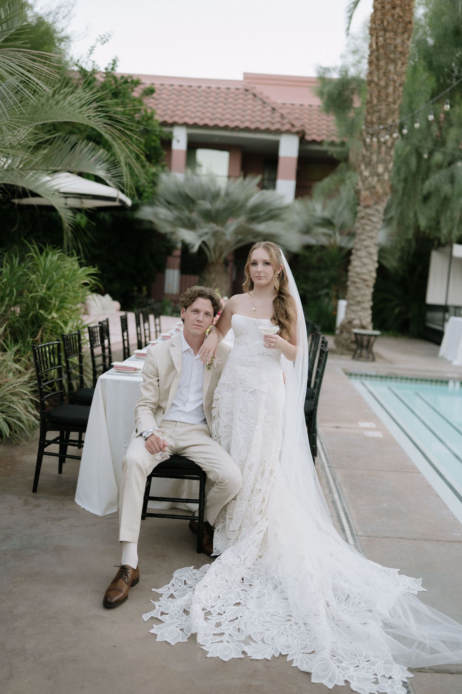Wedding couple in white attire seated by tropical poolside with palm trees.