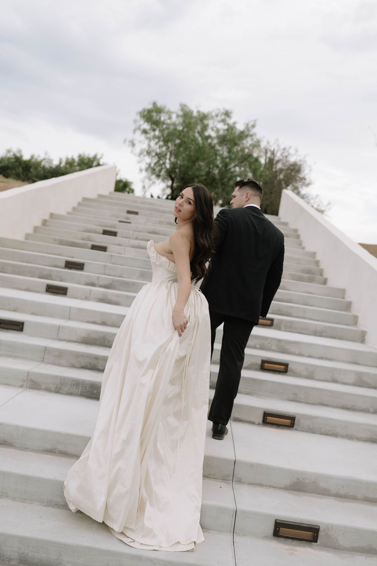 Couple in formal Wedding attire posing dramatically on white concrete steps outdoors.