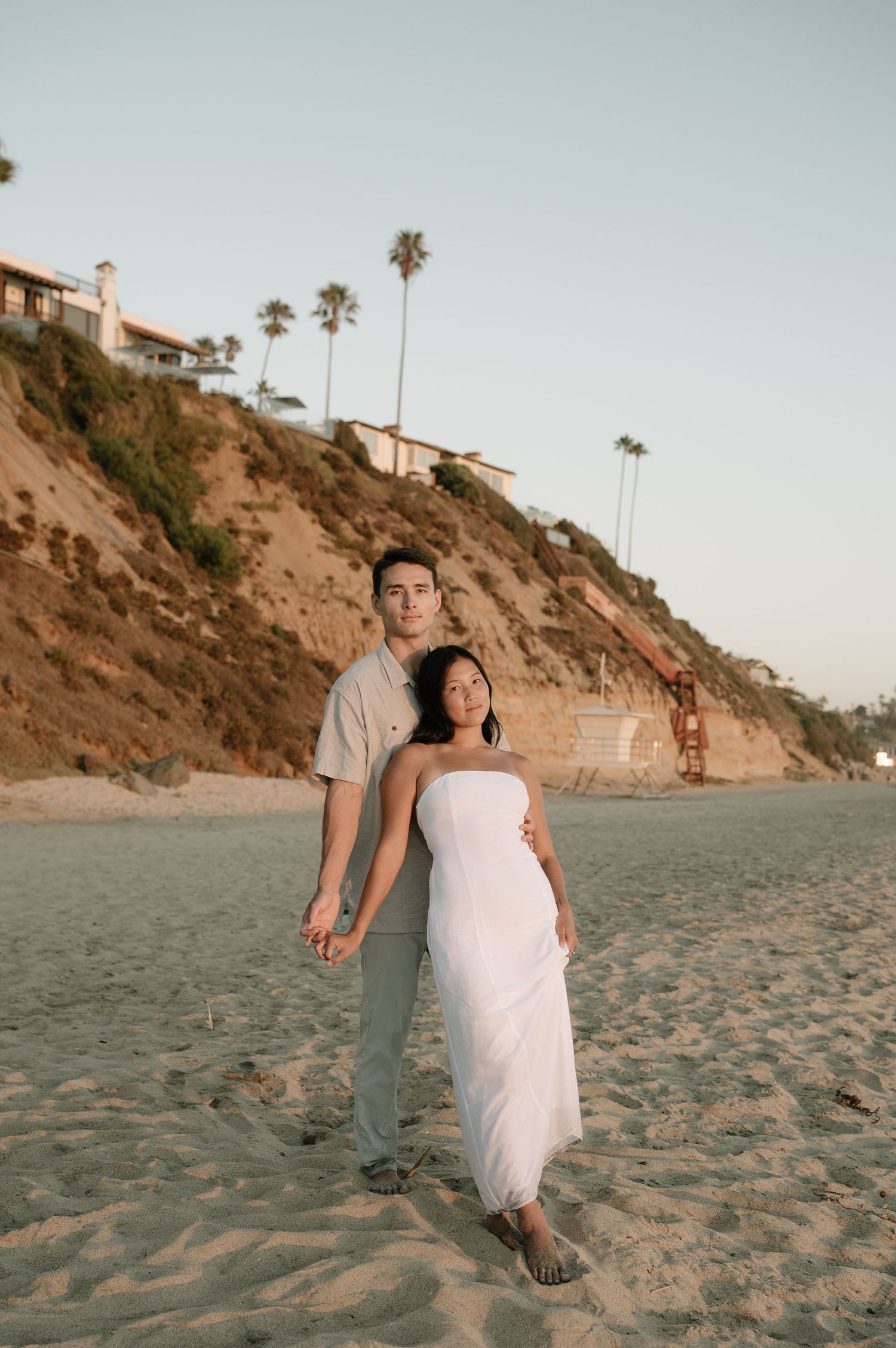A couple walks along a sandy beach with cliffs and palm trees in the background at golden hour.