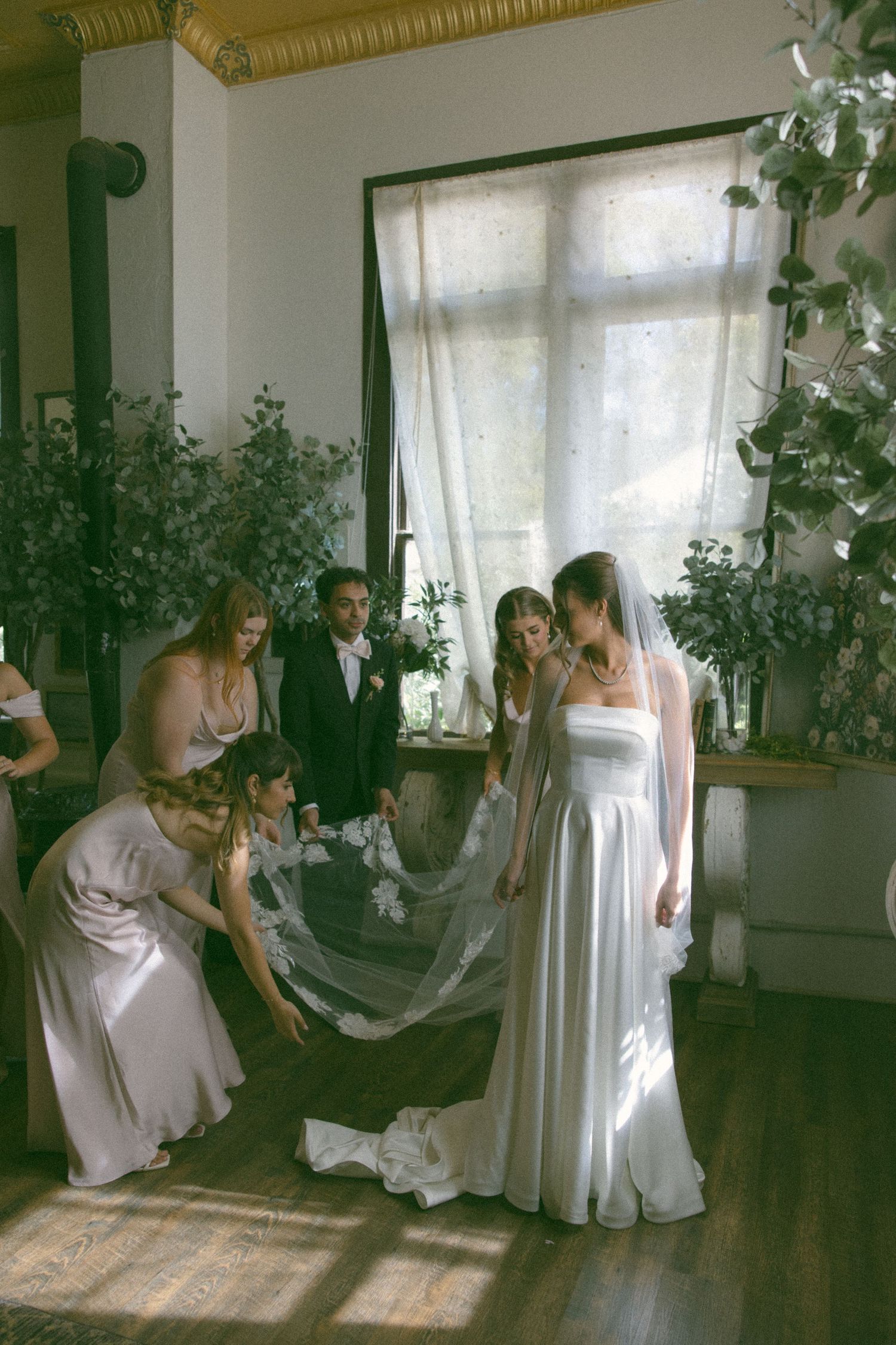 Wedding party helps arrange a white dress in a naturally lit room with indoor plants.