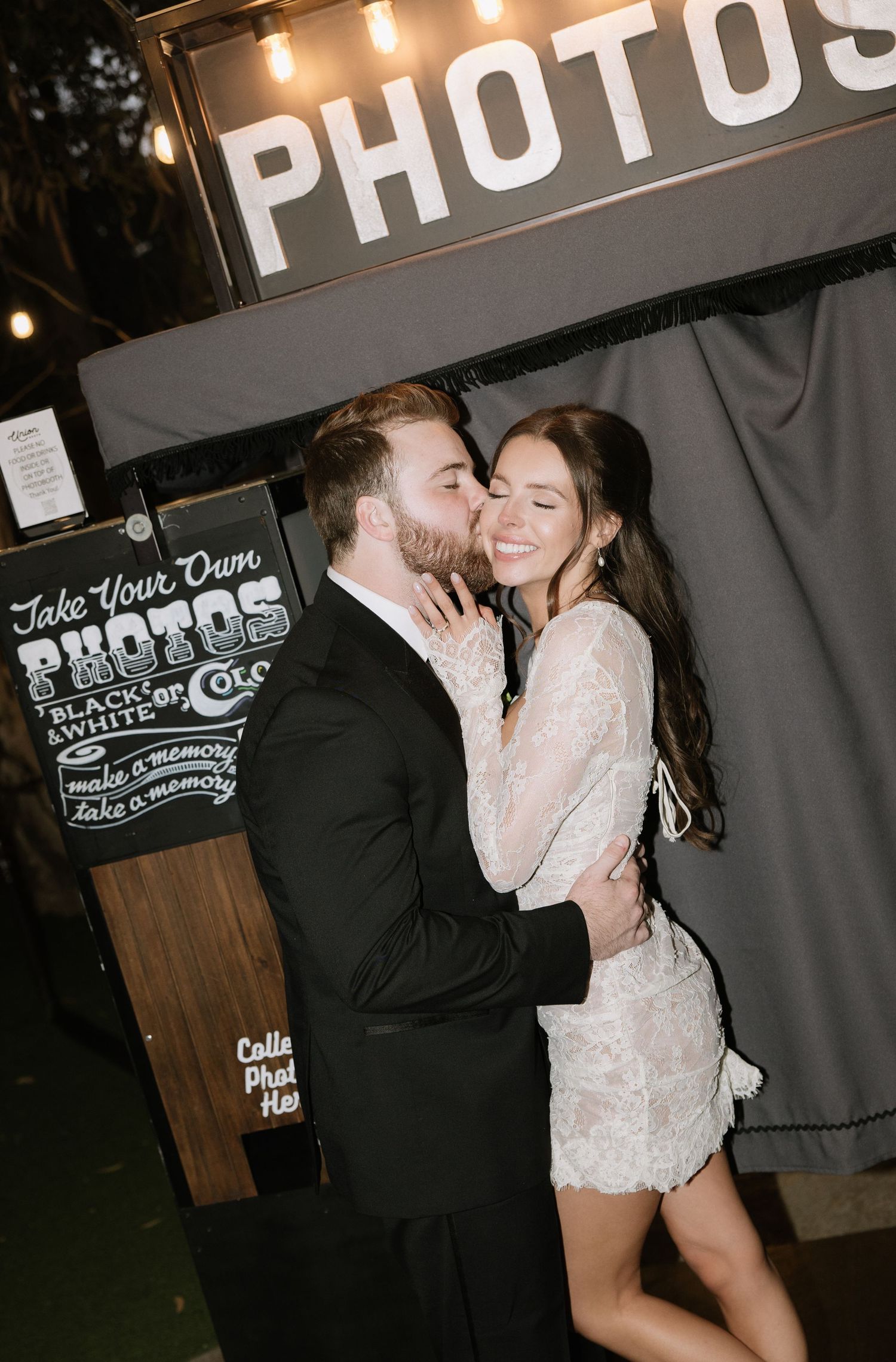 Couple sharing an intimate moment in front of a vintage photo booth sign at night.