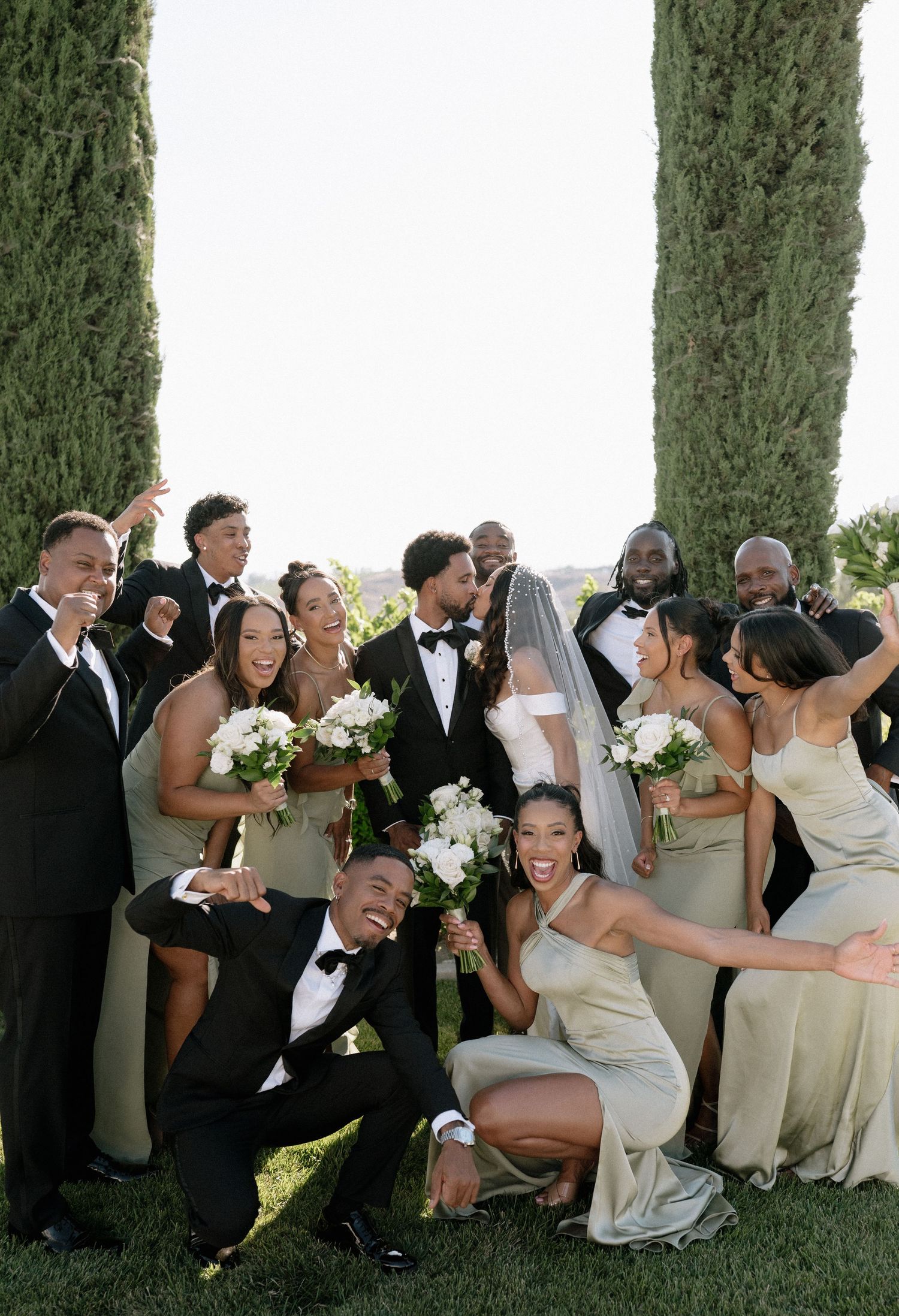 Wedding party poses playfully between cypress trees for group photos.