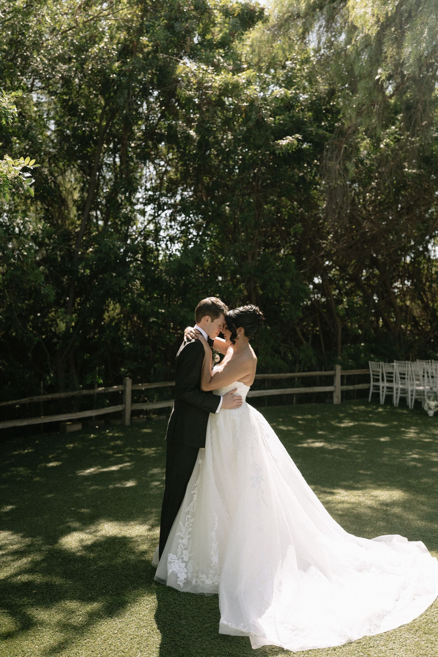 A romantic wedding moment captured outdoors with a flowing white gown against a backdrop of lush greenery and trees.