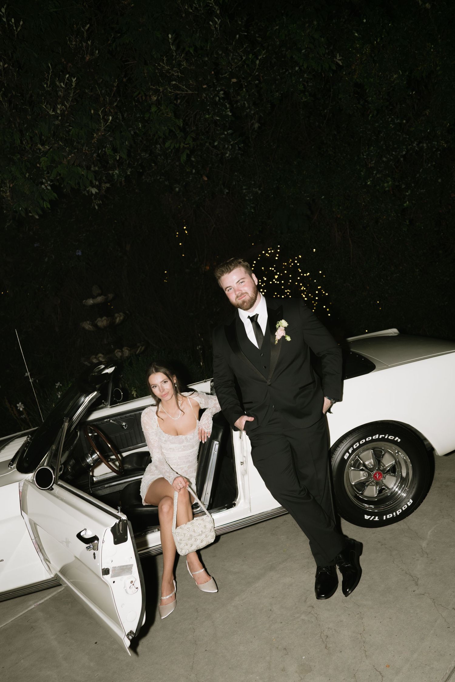Wedding couple sharing a playful moment by a vintage white car with sparklers in background.