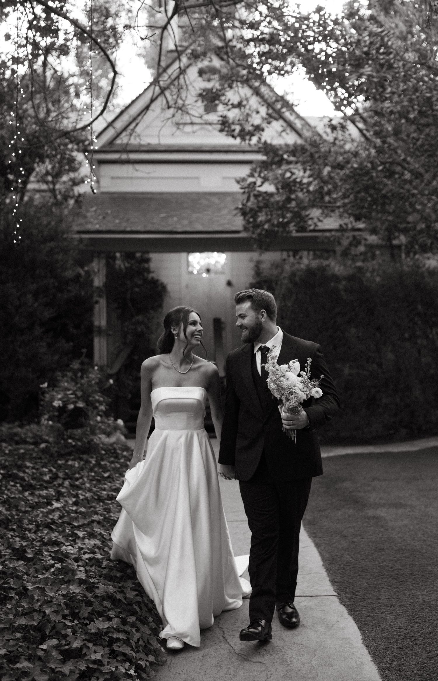 A black and white wedding portrait of a couple walking along a garden path.