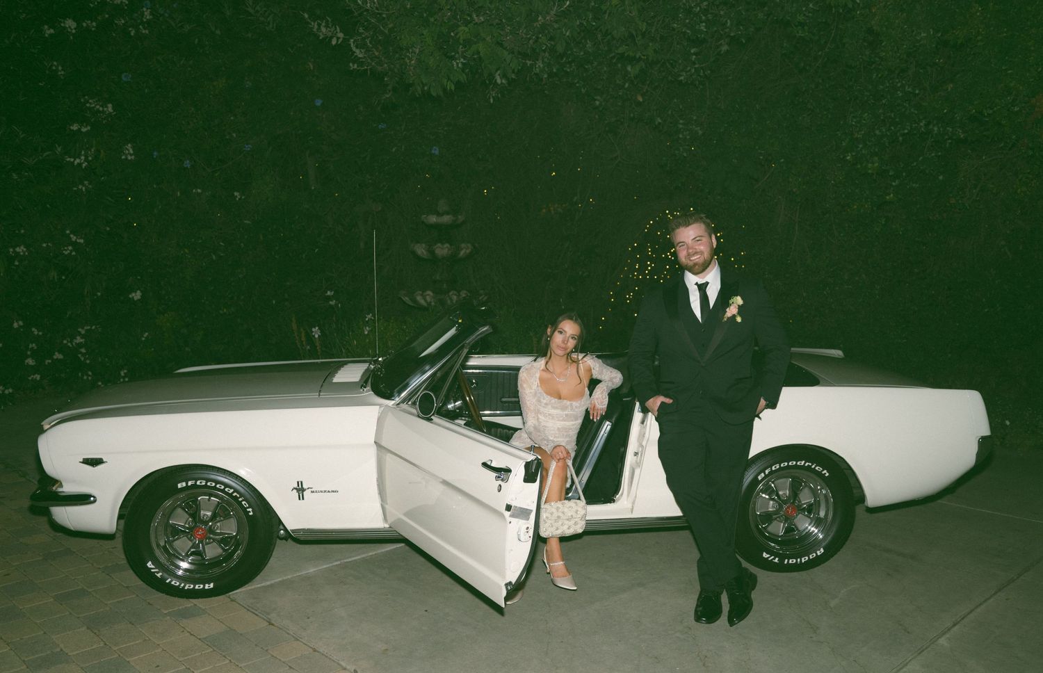 Newlyweds posing with a classic white Mustang car at night.