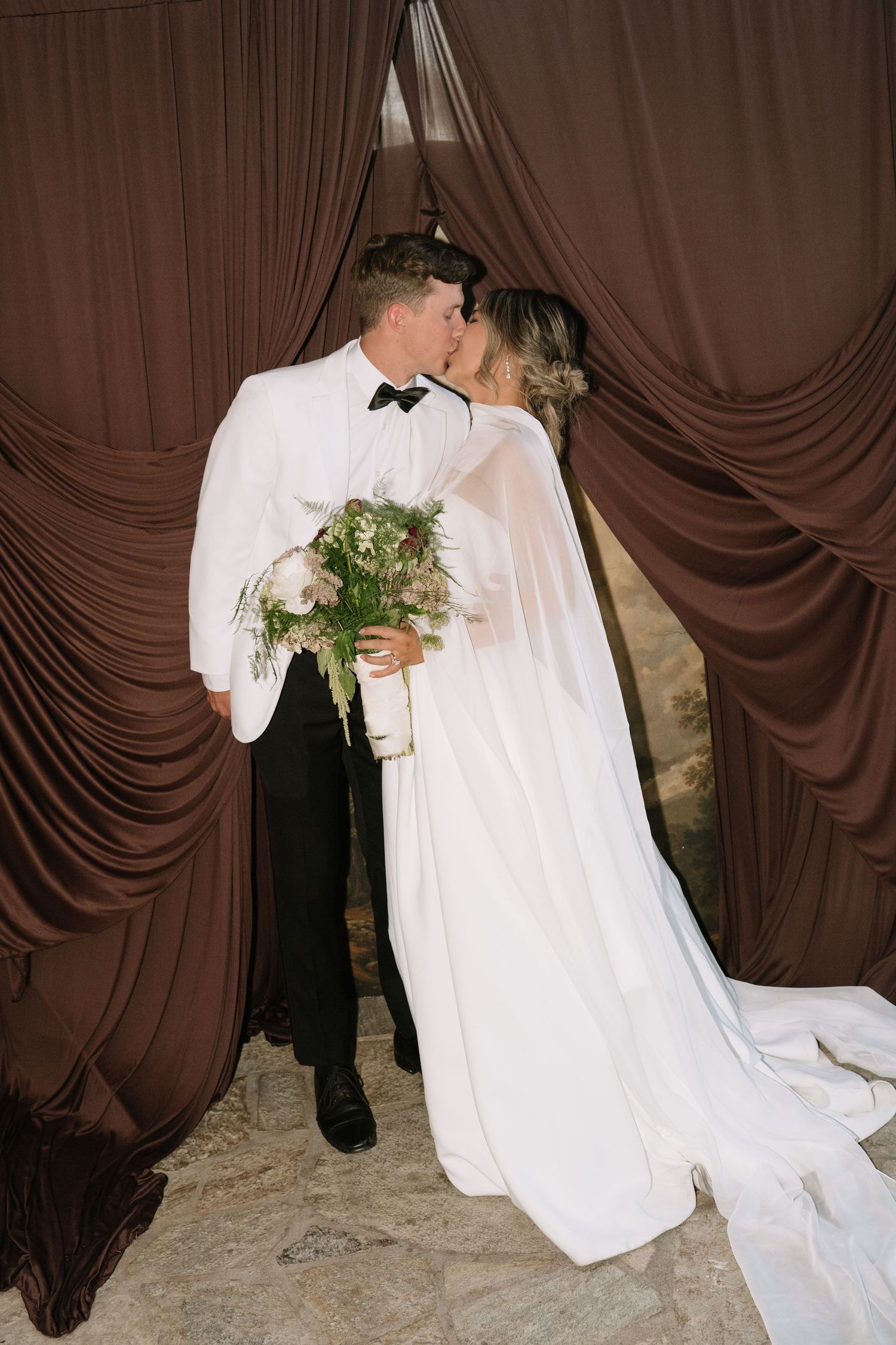 Couple in wedding attire sharing an intimate moment against brown draped backdrop.