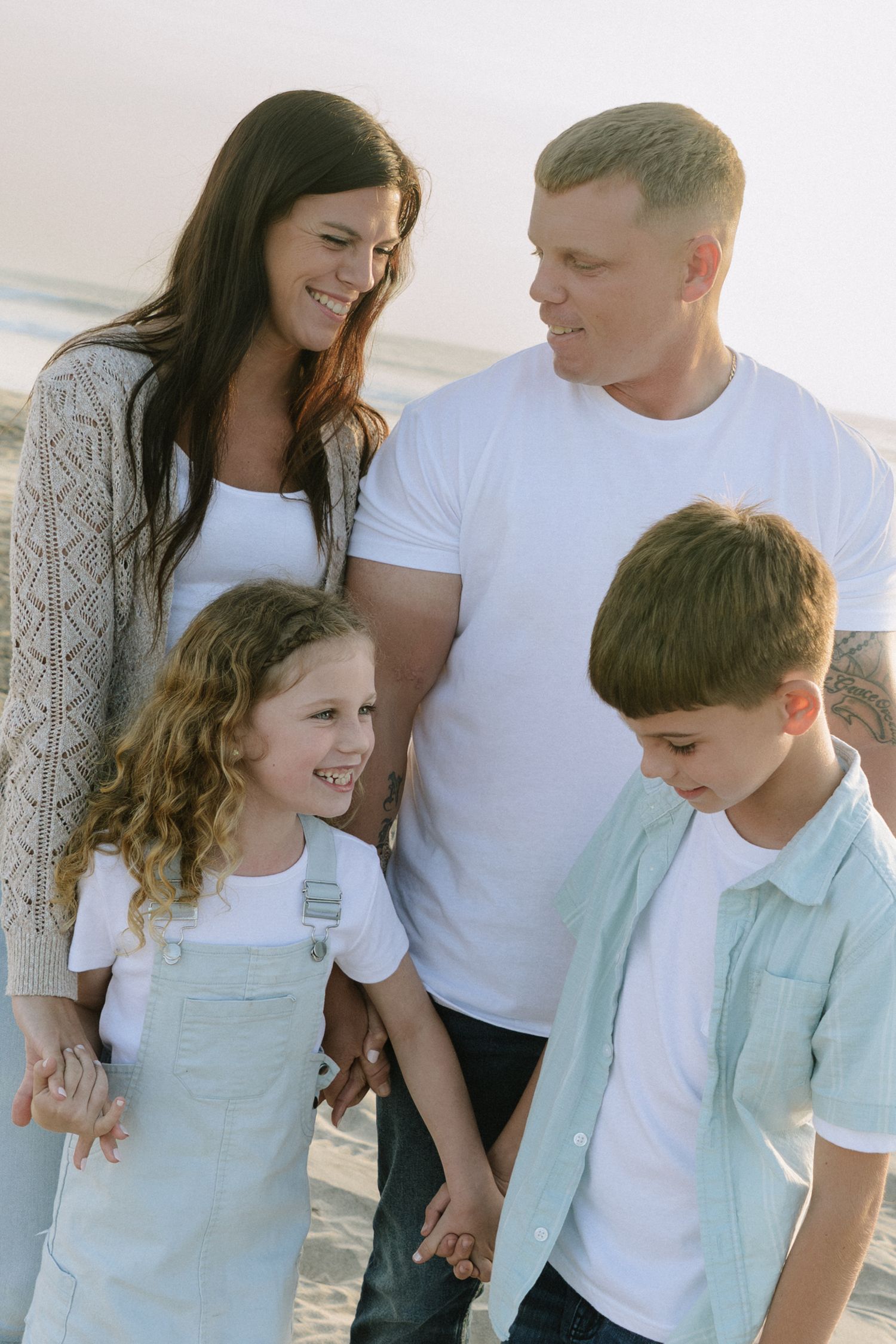 A family of four wearing coordinated white and mint outfits shares laughs on the beach.
