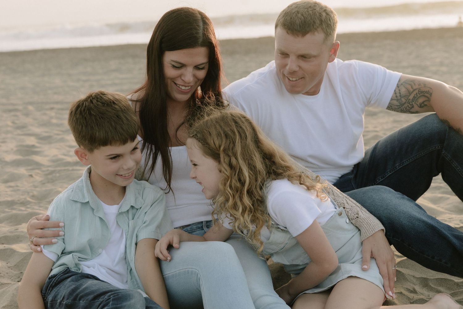 Family members cuddle and laugh together while sitting on beach sand.