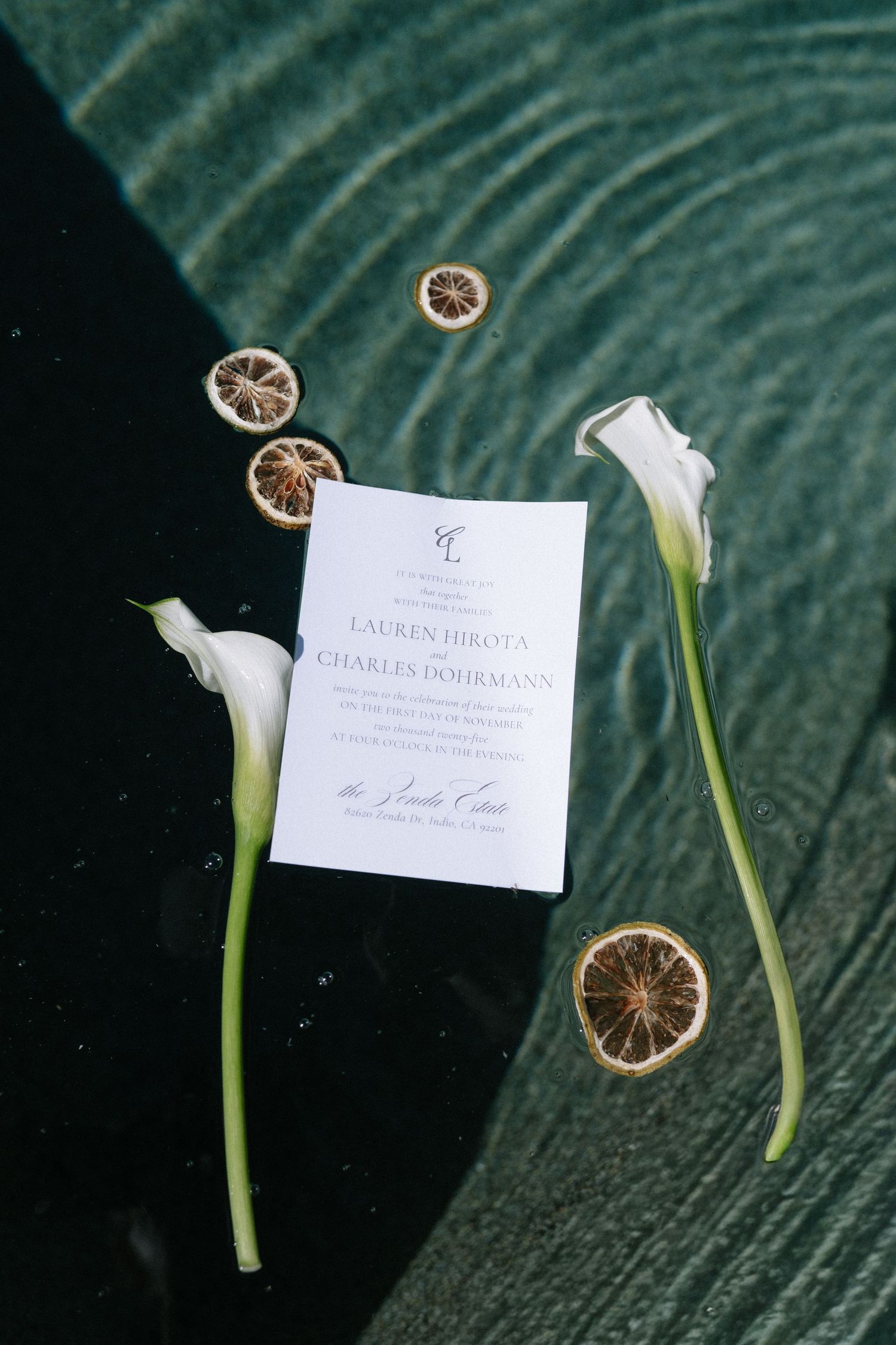 Wedding invitation styled with white calla lilies and dried citrus slices on a dark green surface.