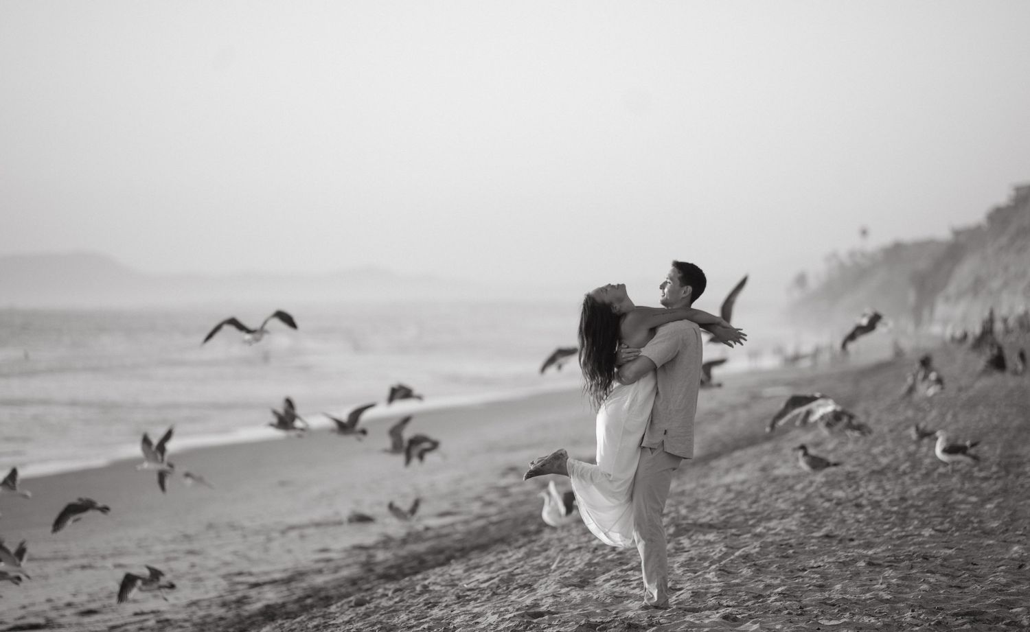 A couple in white clothing chases seagulls on a misty beach in black and white.