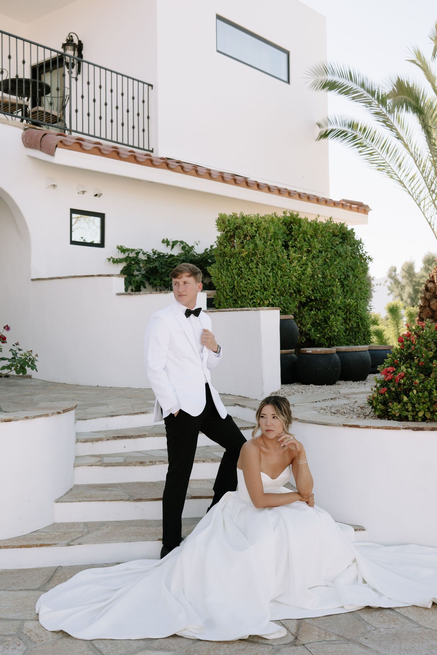 Wedding couple posing on white steps of Mediterranean-style villa with palm trees.