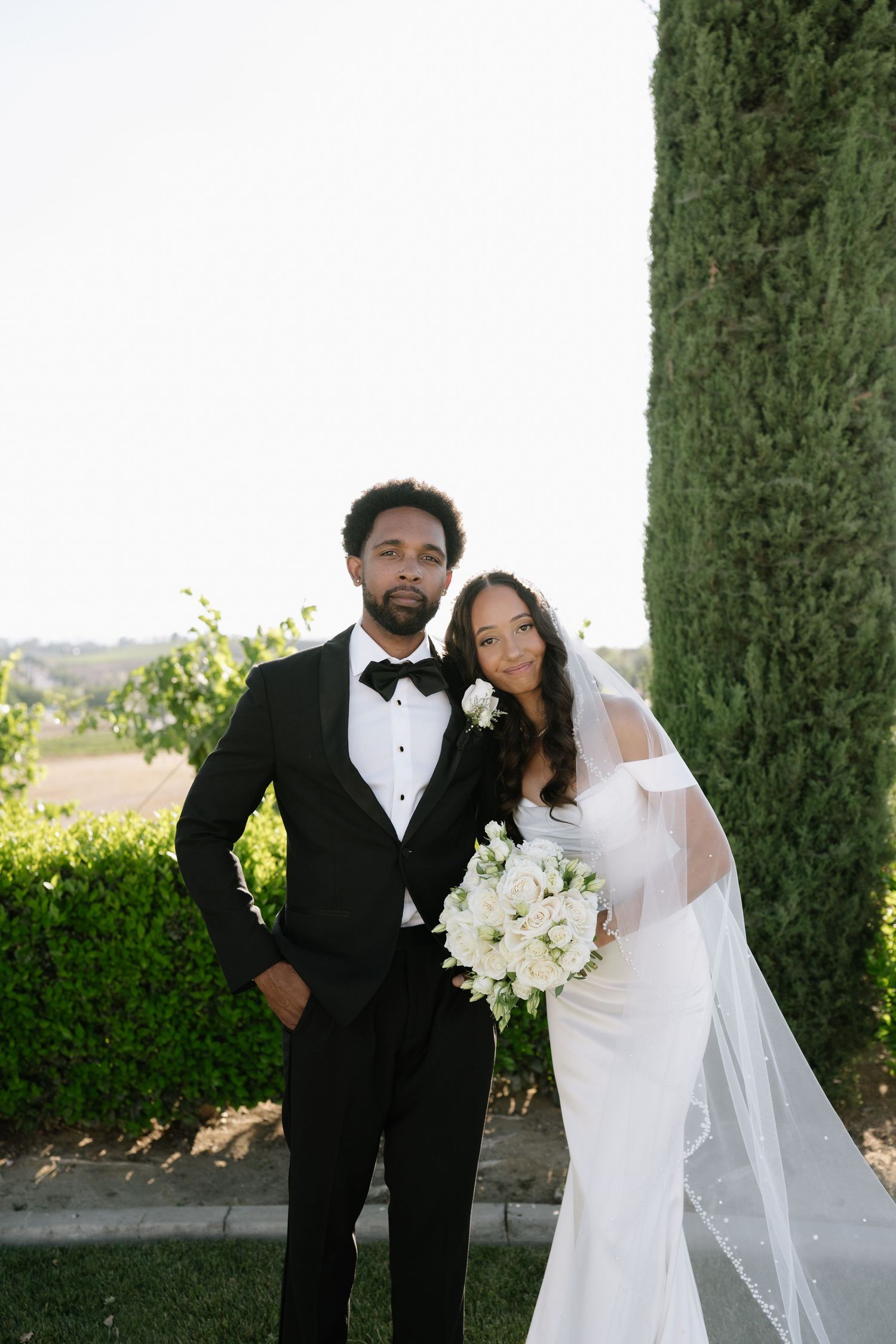 Wedding portrait of couple in formal attire standing beside cypress tree with scenic backdrop.