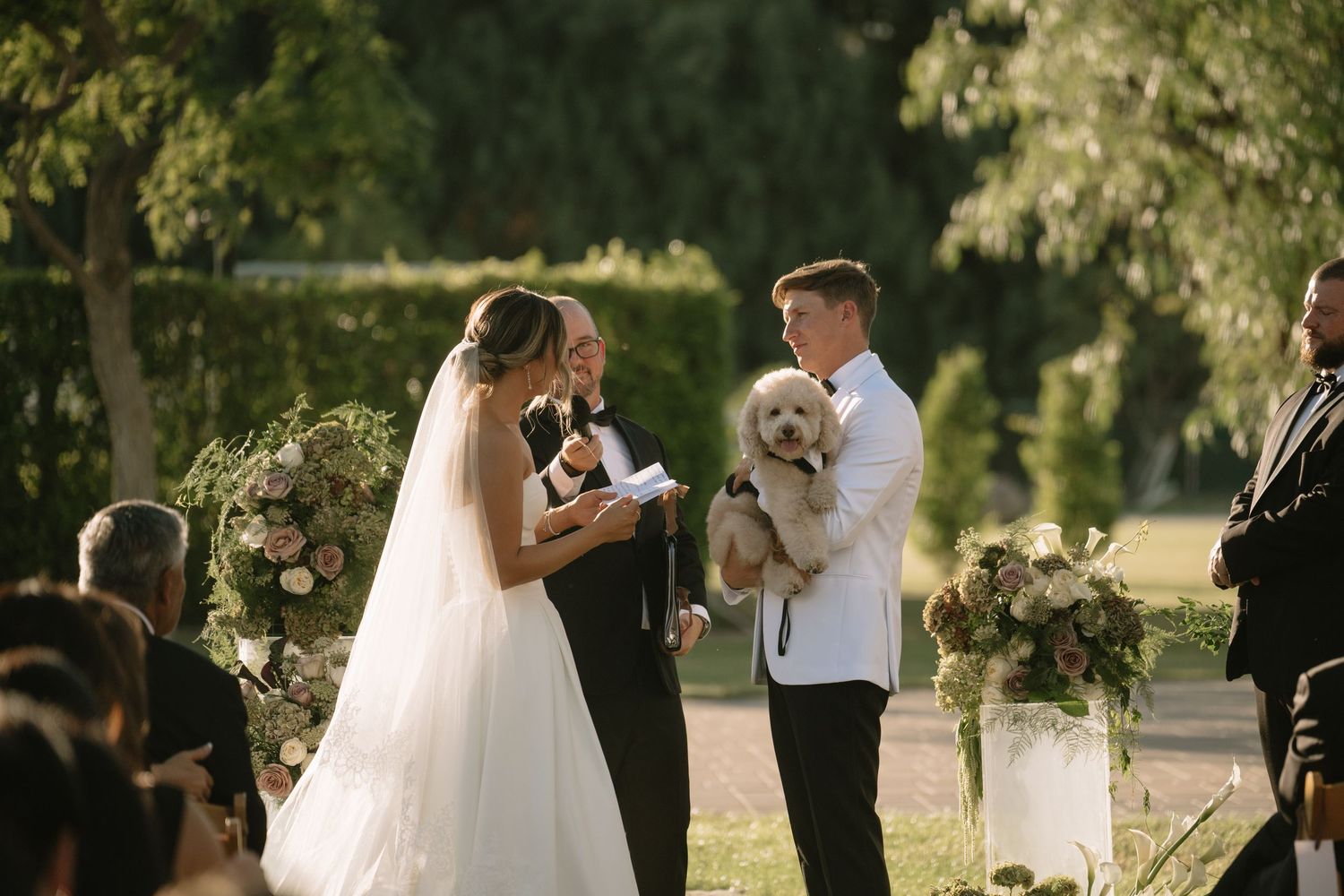 Outdoor wedding ceremony with bridal party and guests gathered under trees.