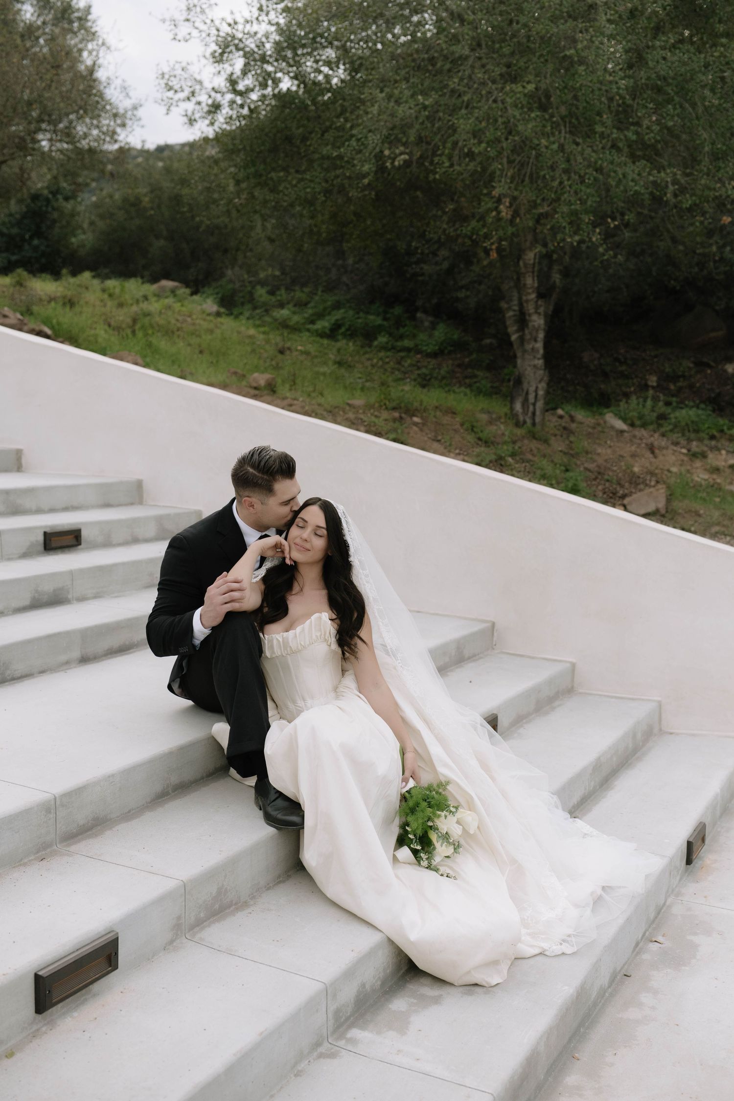 Romantic couple in wedding attire lounging on white outdoor steps.