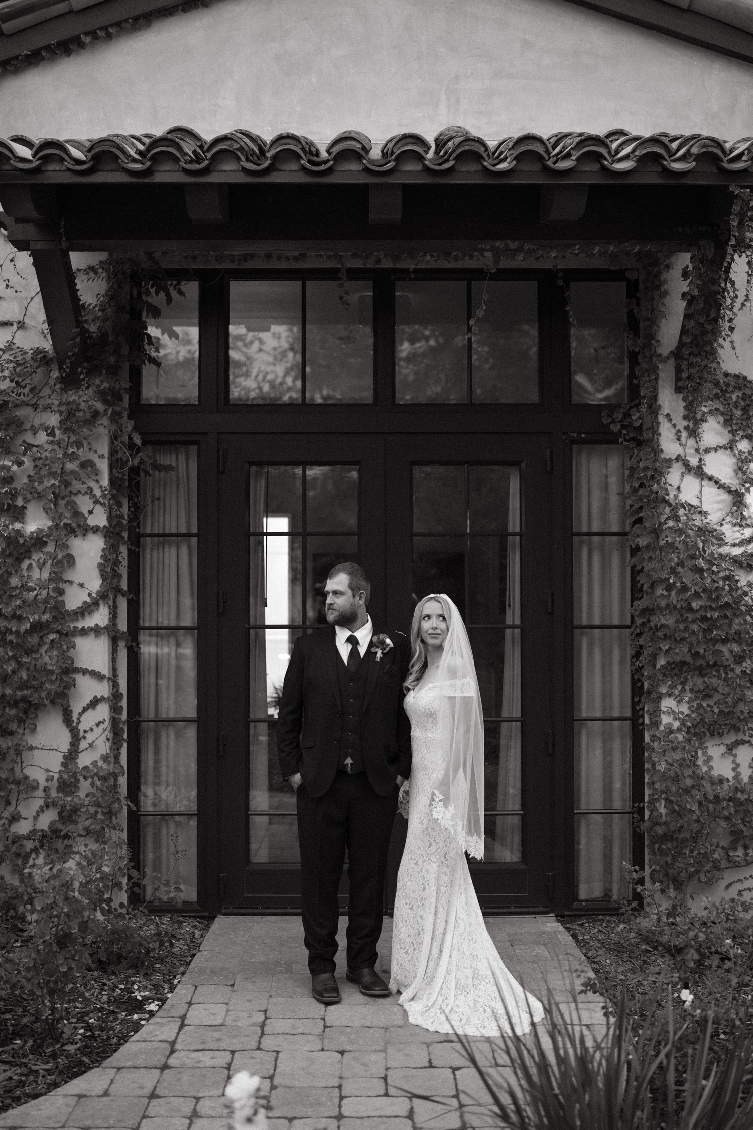 A couple in wedding attire poses in front of a Spanish-style building with arched doorways.