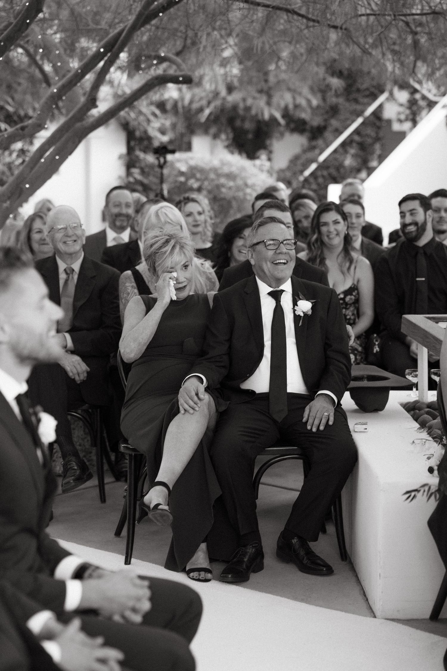 Wedding ceremony guests laugh and smile during an outdoor celebration.