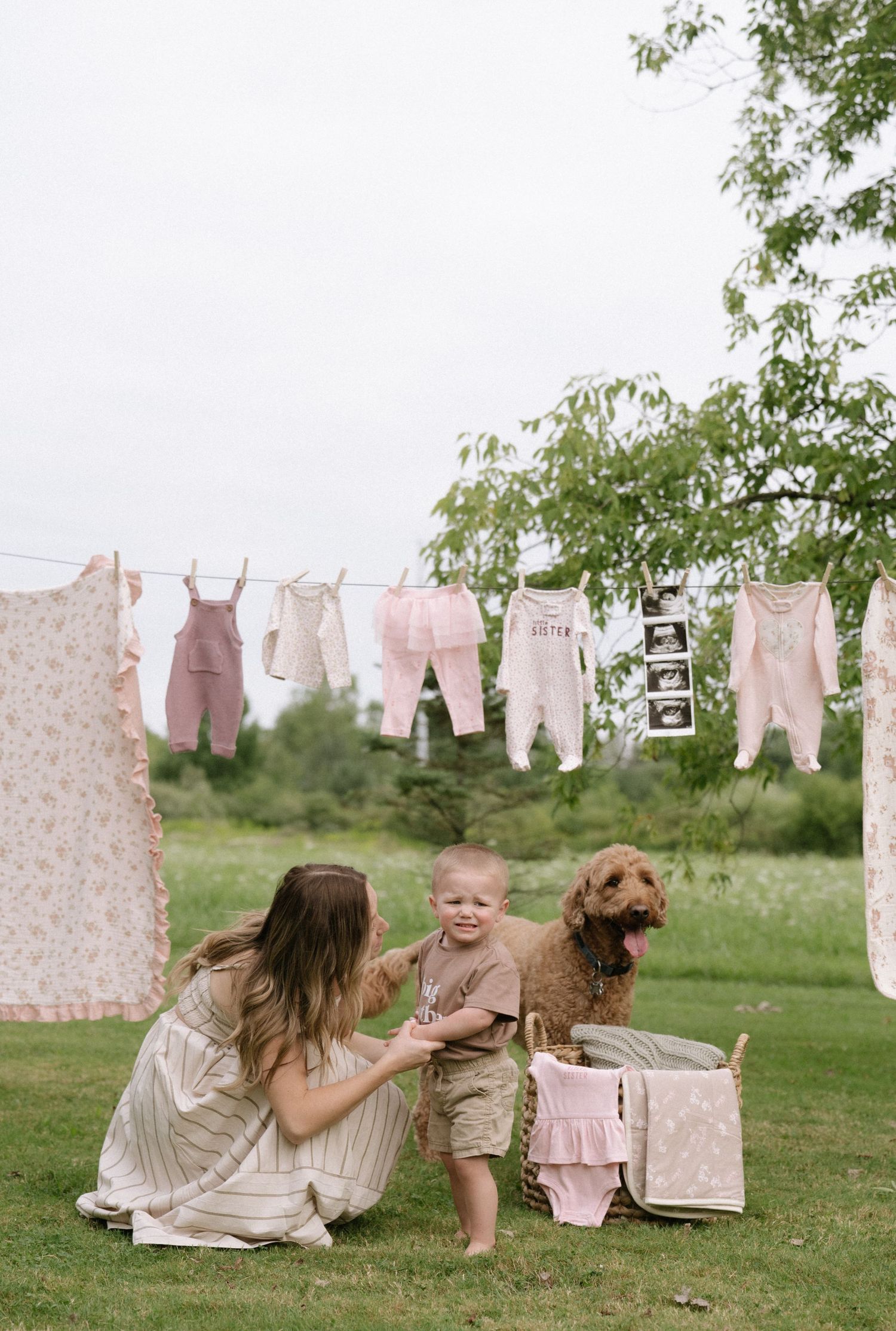 Baby clothes and blankets hanging on a clothesline in a sunny garden with a dog present.