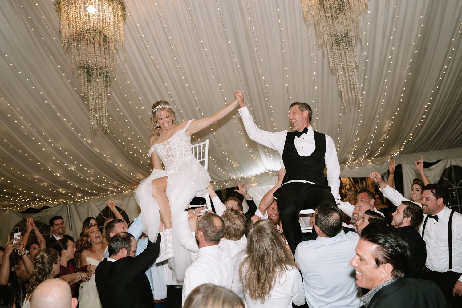 Wedding reception guests dancing and celebrating under crystal chandeliers in an elegant venue.