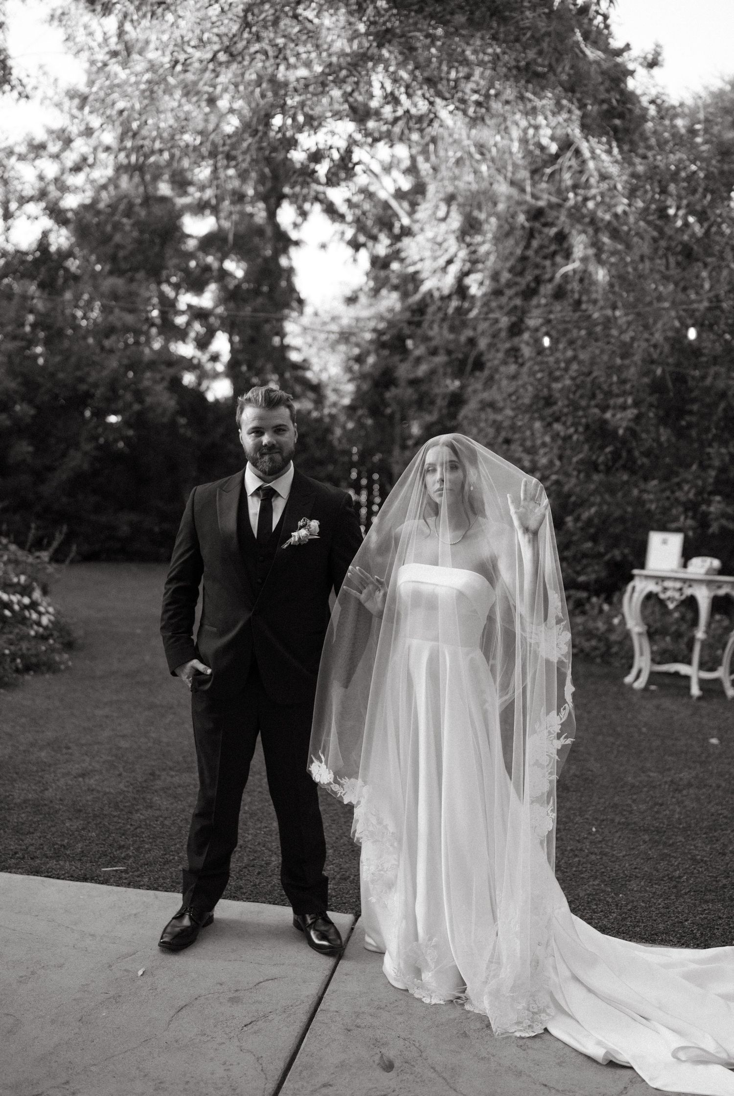 A black and white portrait of a couple in wedding attire standing on a garden path.
